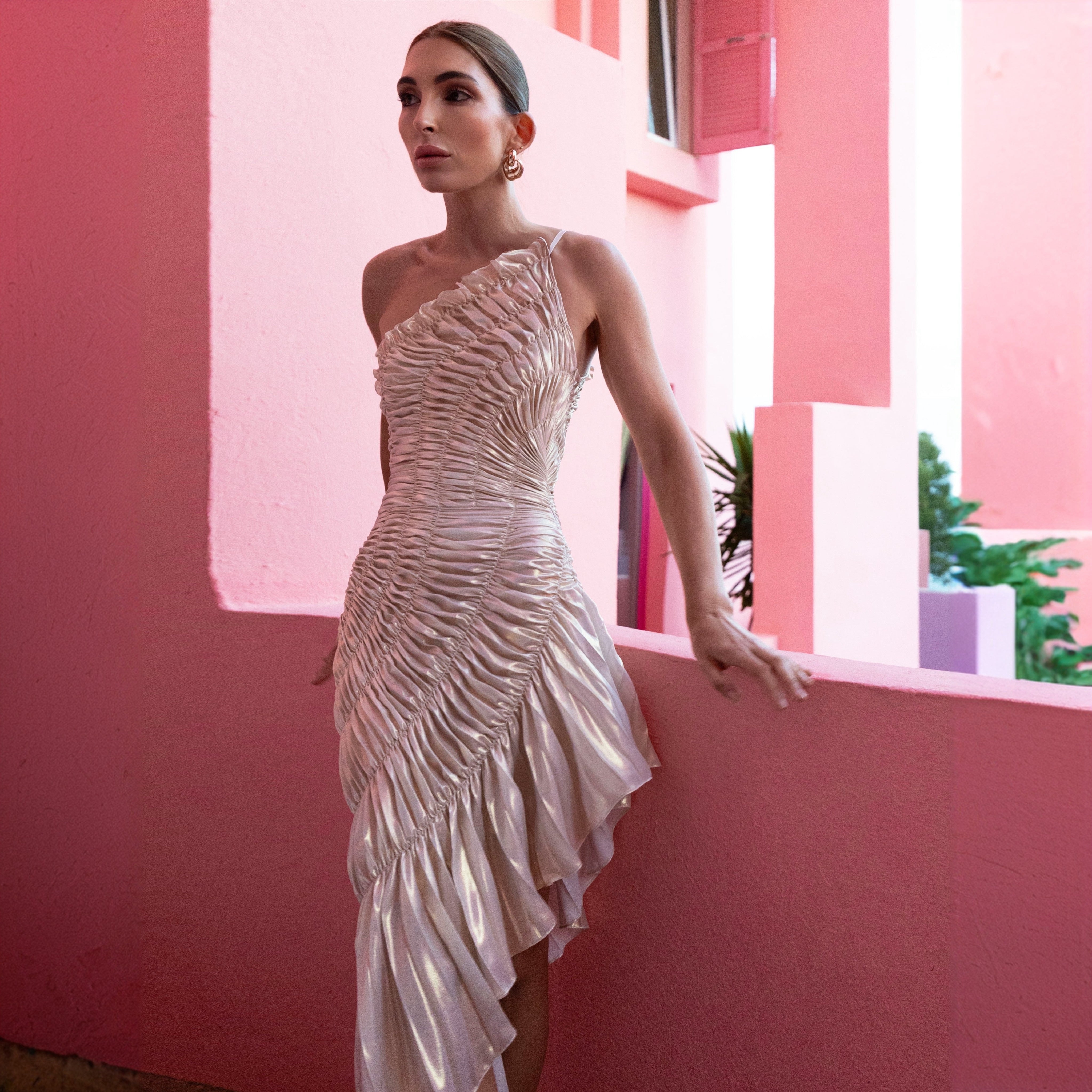 Woman in a silver one-shoulder dress standing against a pink wall with a window.