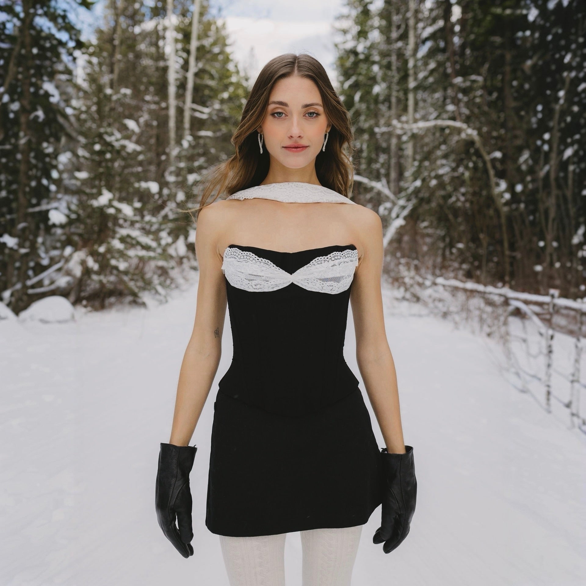 Woman in a black dress with white lace details standing in a snowy forest.