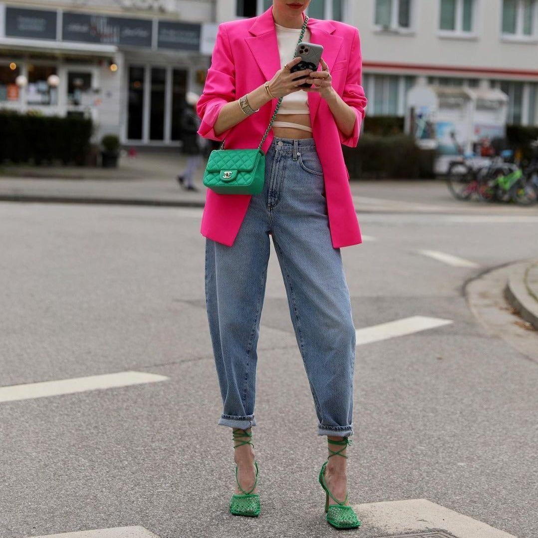 Person wearing a bright pink blazer and blue jeans, holding a green handbag on an urban street.