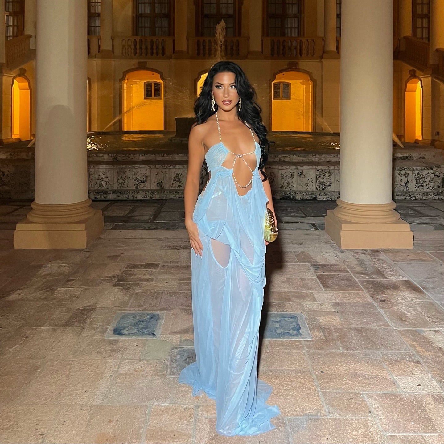 Woman in a light blue dress standing in an elegant indoor setting with columns and warm lighting.