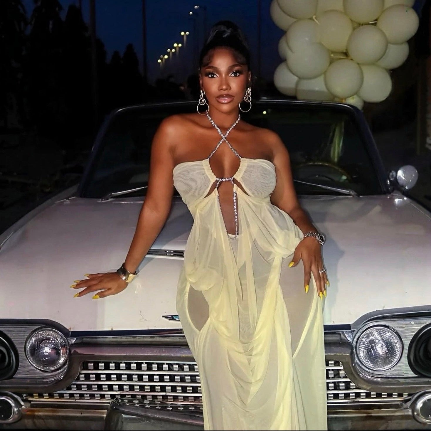 Woman in a yellow dress standing in front of a classic car with balloons in the background