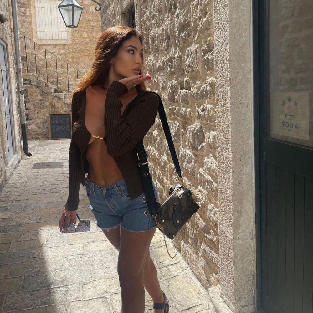 Woman in a brown cardigan and denim shorts standing against a stone wall.