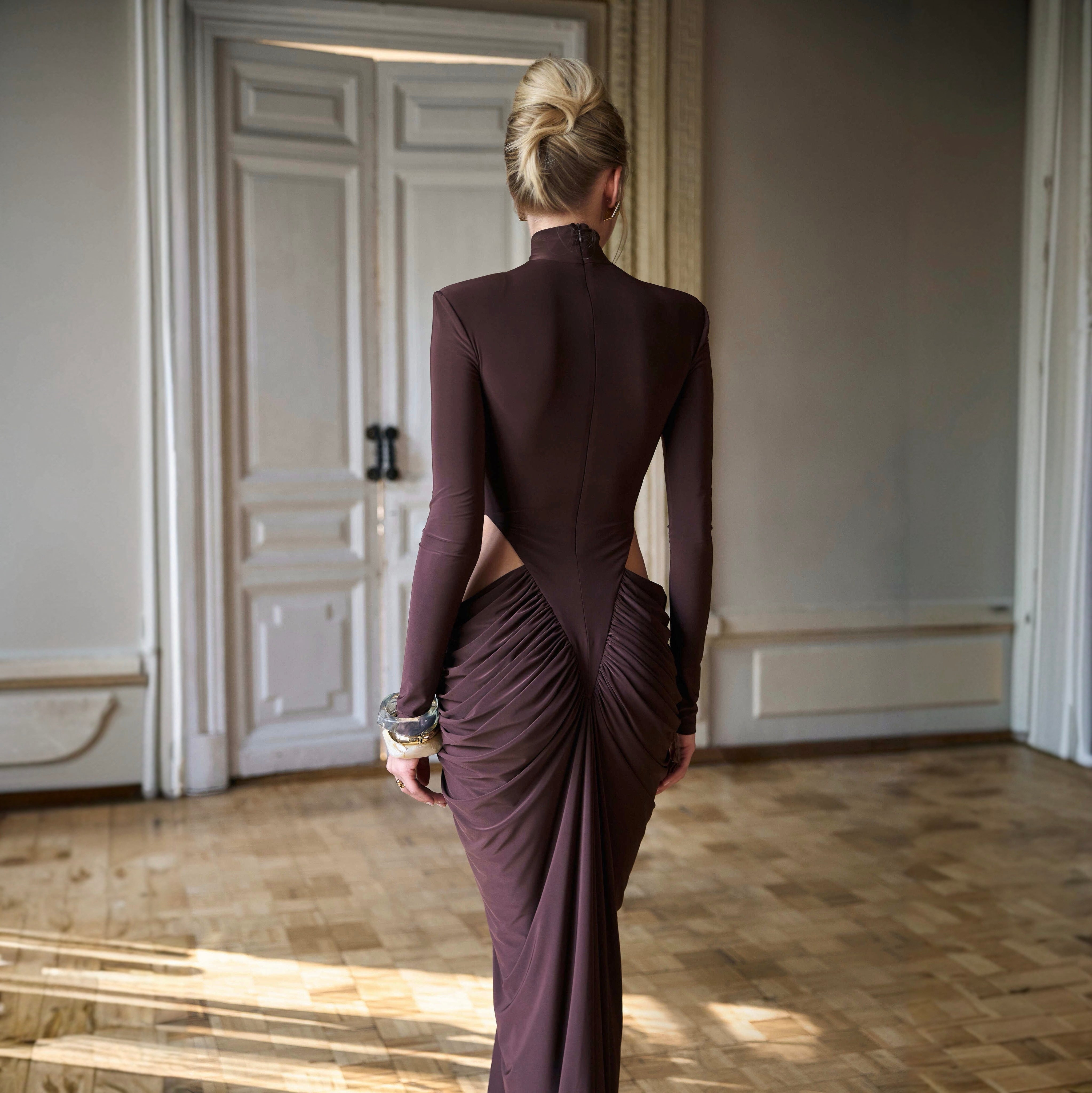 Woman in a long, dark brown dress with an open back standing in a room with wooden floors and white walls.