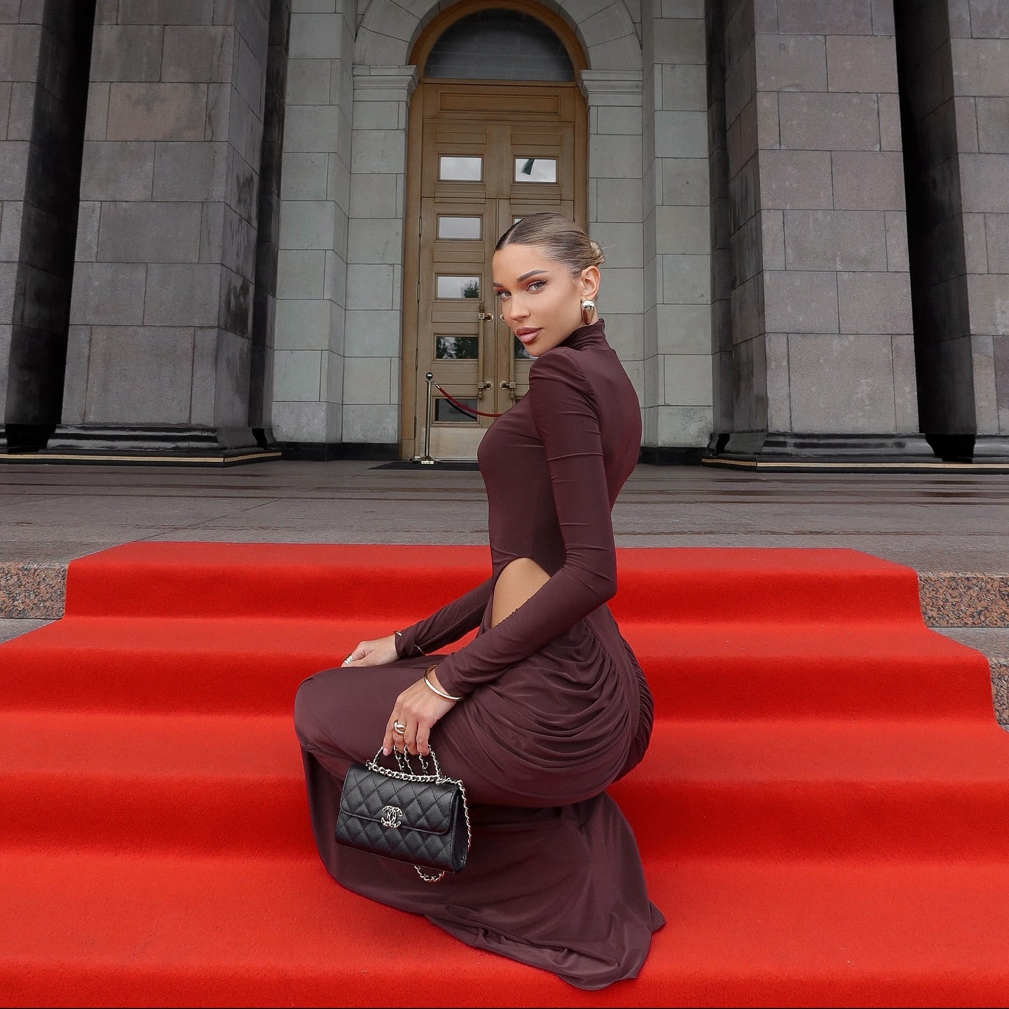 Woman in a burgundy dress sitting on red steps in front of a stone building.
