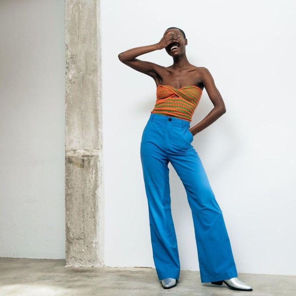Woman wearing an orange top and blue pants against a white wall.