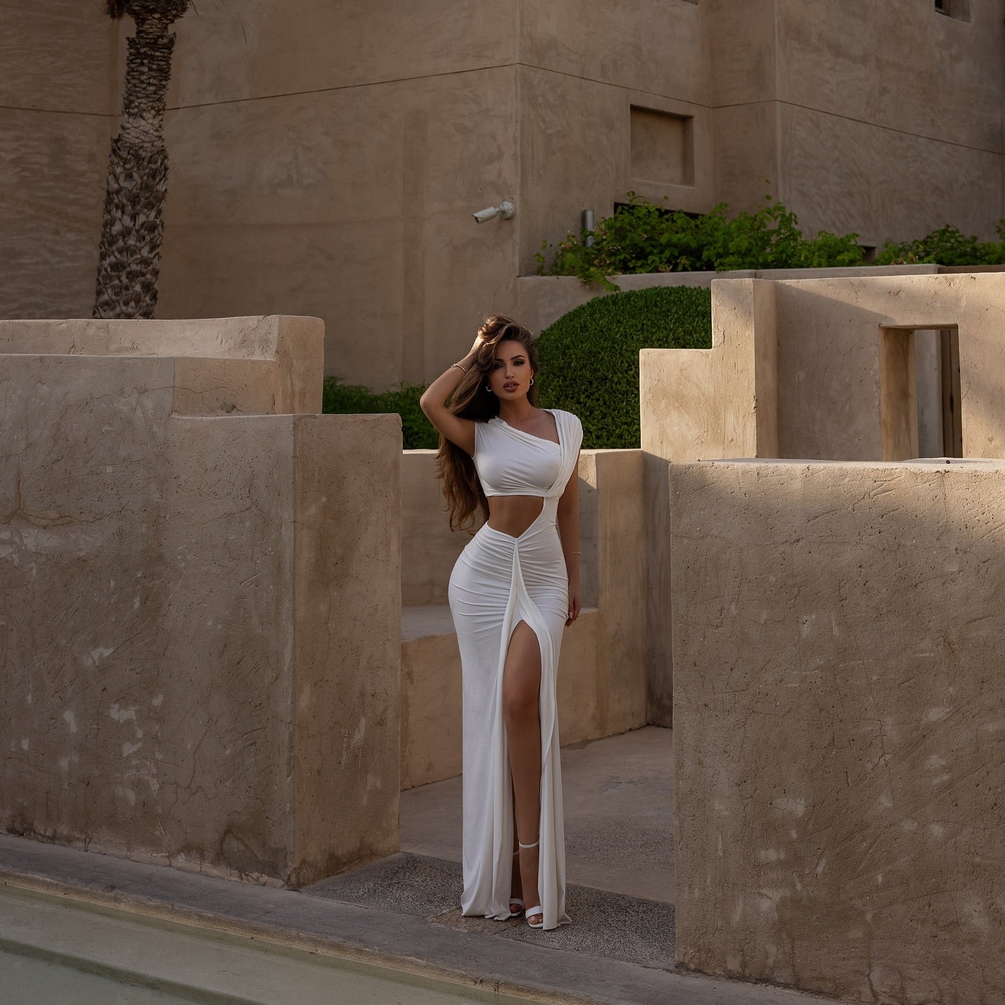 Woman in a white outfit standing in front of a beige building with palm trees.