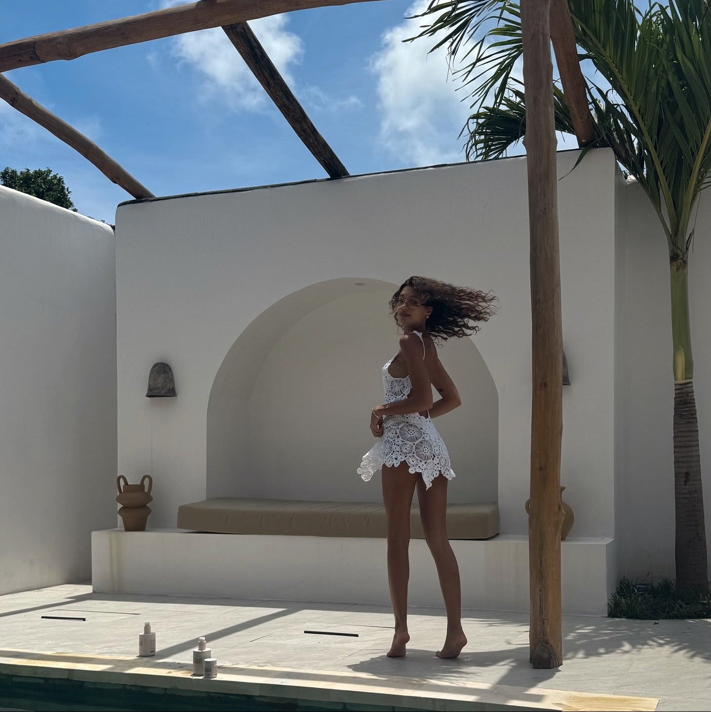 Woman in a white outfit standing under a wooden pergola with a clear blue sky and palm trees in the background.