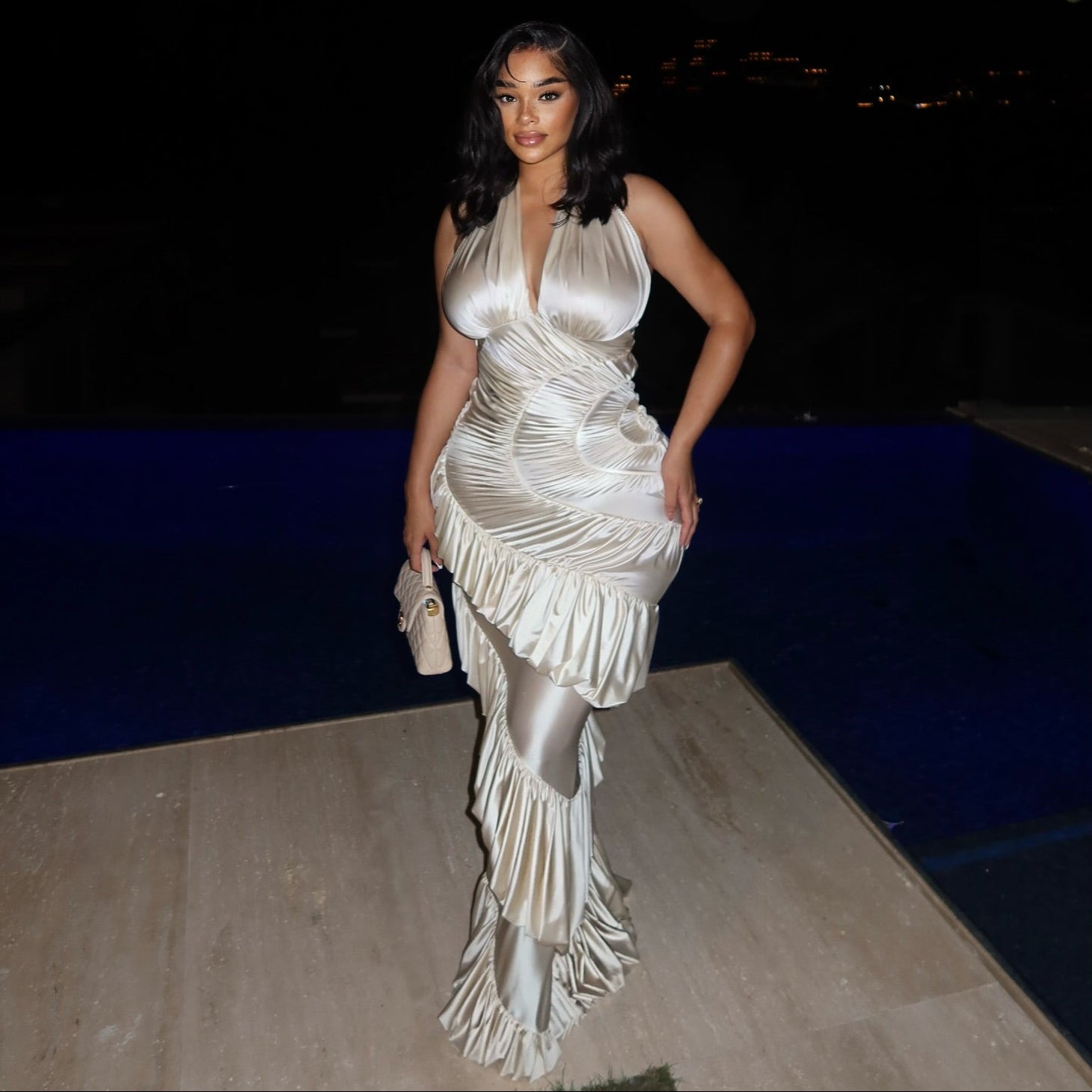 Woman in a white evening gown standing by a pool at night.