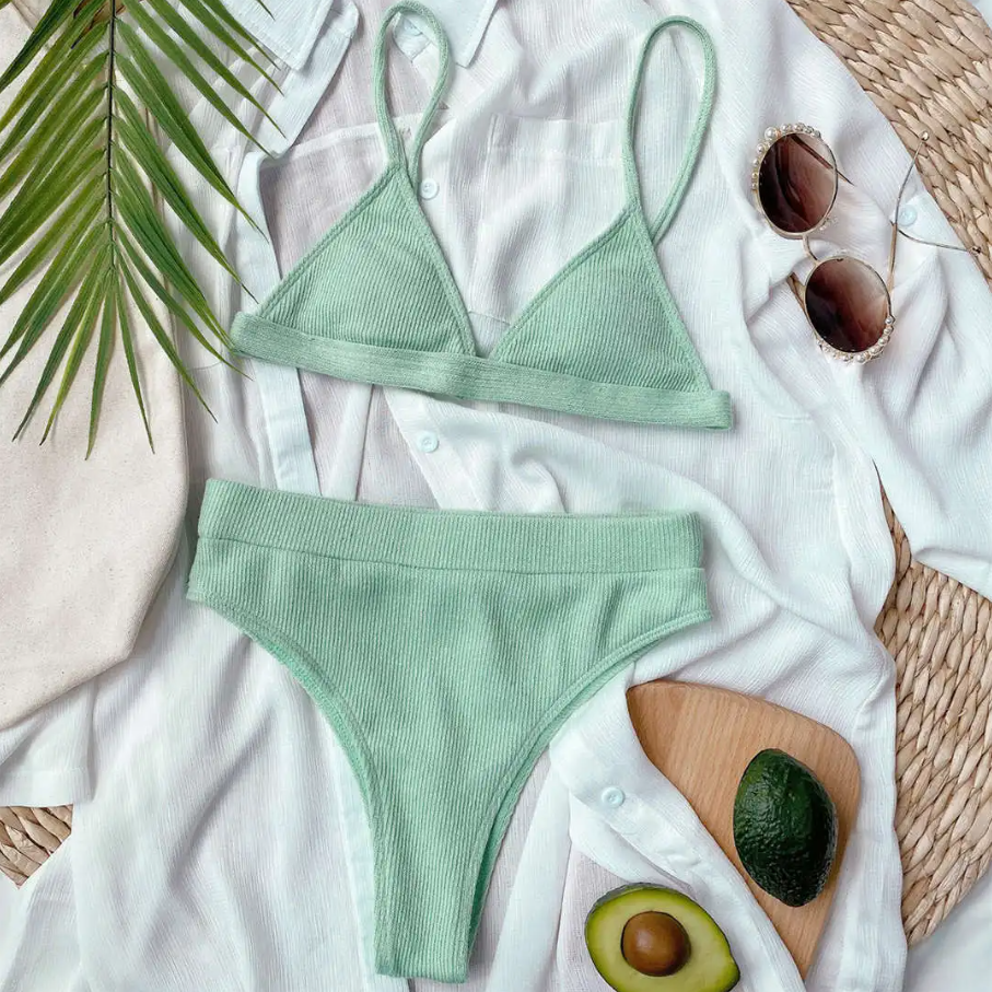 Green bikini set on a white surface with sunglasses, avocado, and palm leaves.