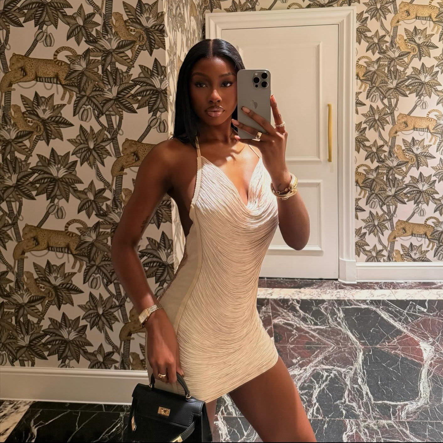 Woman taking a mirror selfie in a beige dress with floral wallpaper in the background