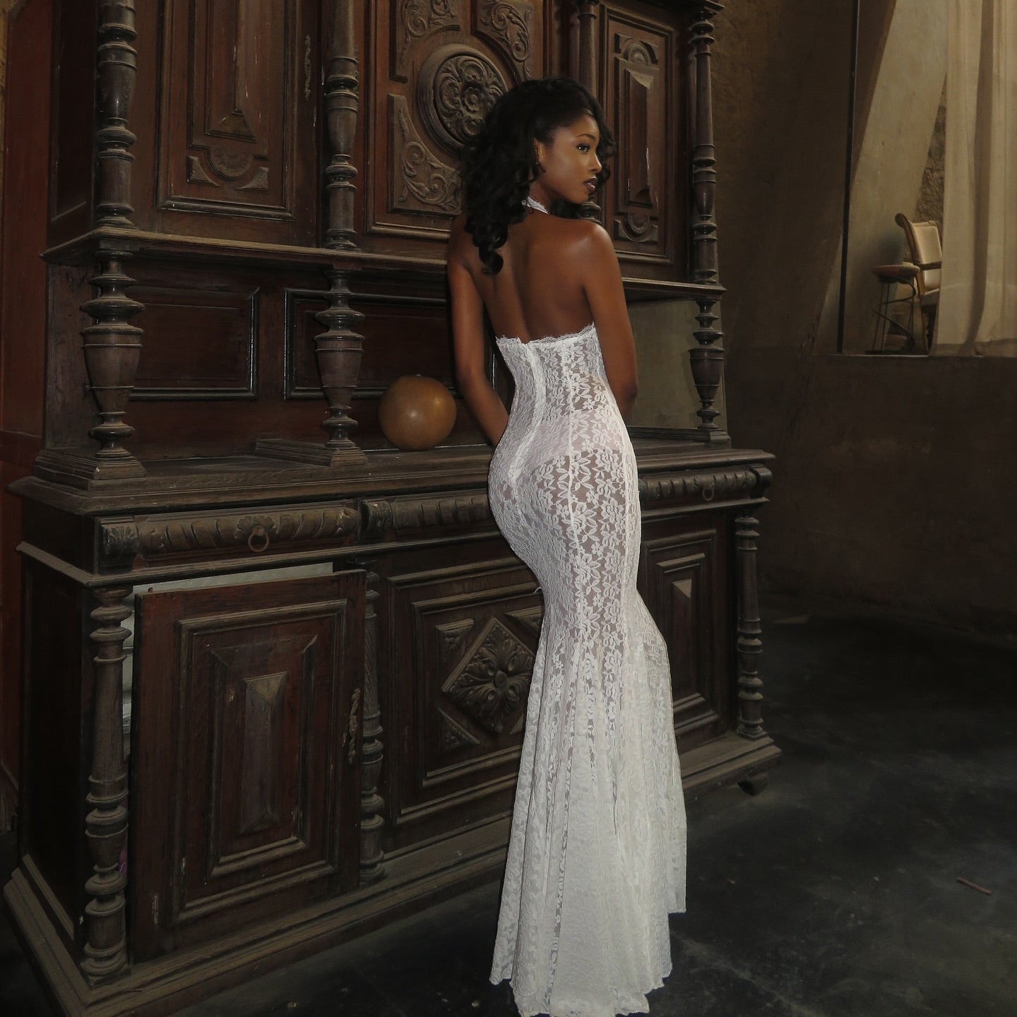 Woman in a white lace dress standing in front of an ornate wooden cabinet.