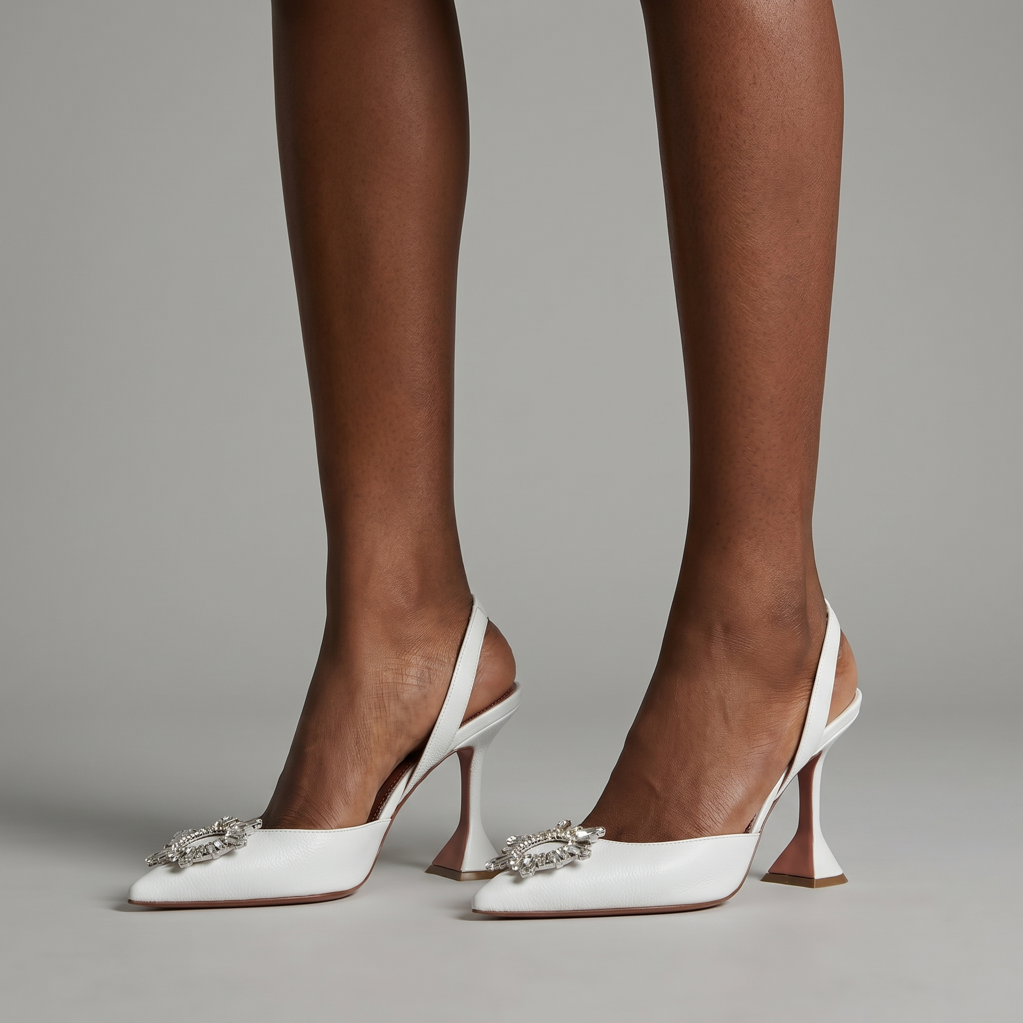 White high-heeled shoes with embellishments worn by a person on a gray background