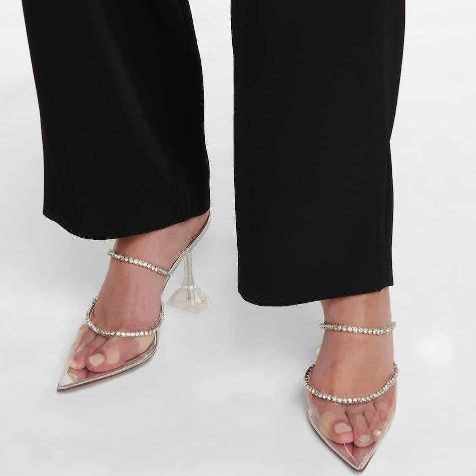Clear high-heeled shoes with embellishments worn with black pants on a white background