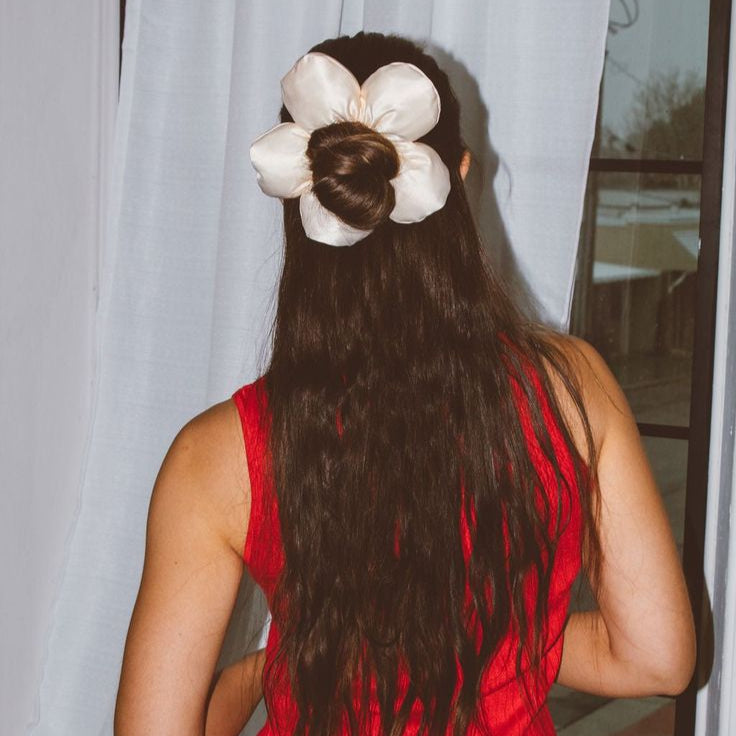 Person with long hair tied up with a large white flower, wearing a red sleeveless top, standing in front of a light curtain.