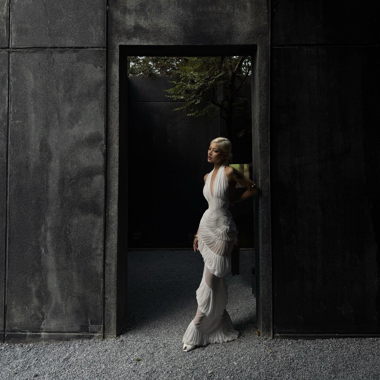 Woman in a white dress standing in a dark, modern architectural setting with concrete walls and trees in the background.
