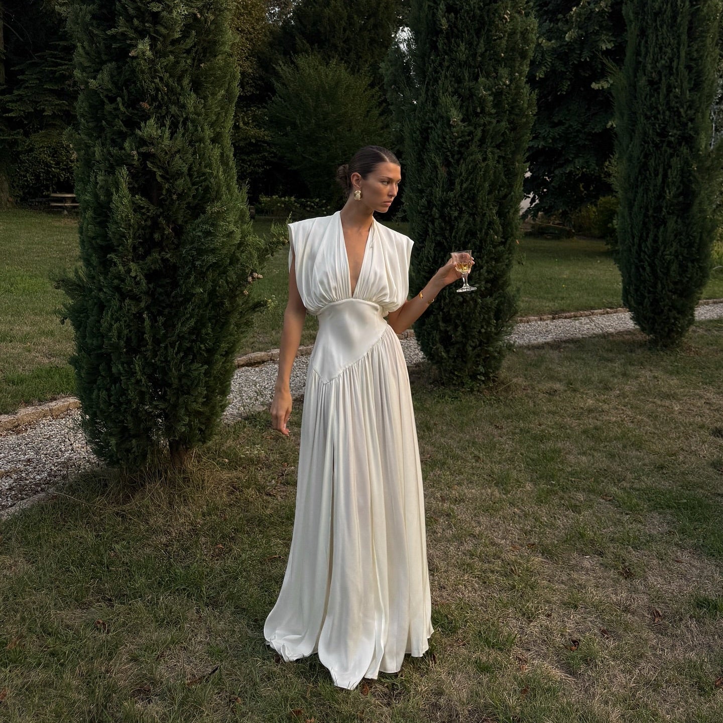 Woman in a white dress standing in a garden with tall green trees.