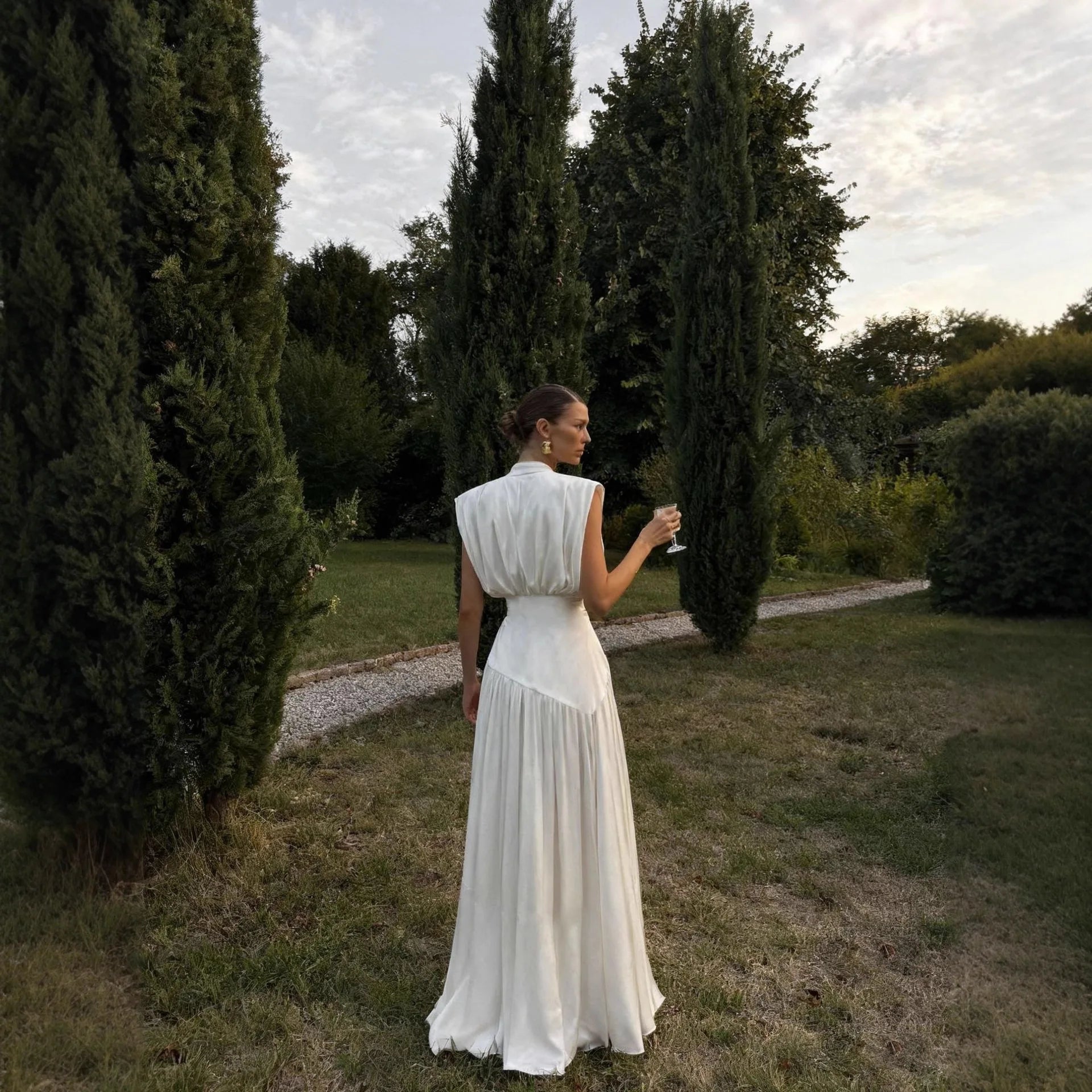Woman in a white dress standing in a garden holding a glass.