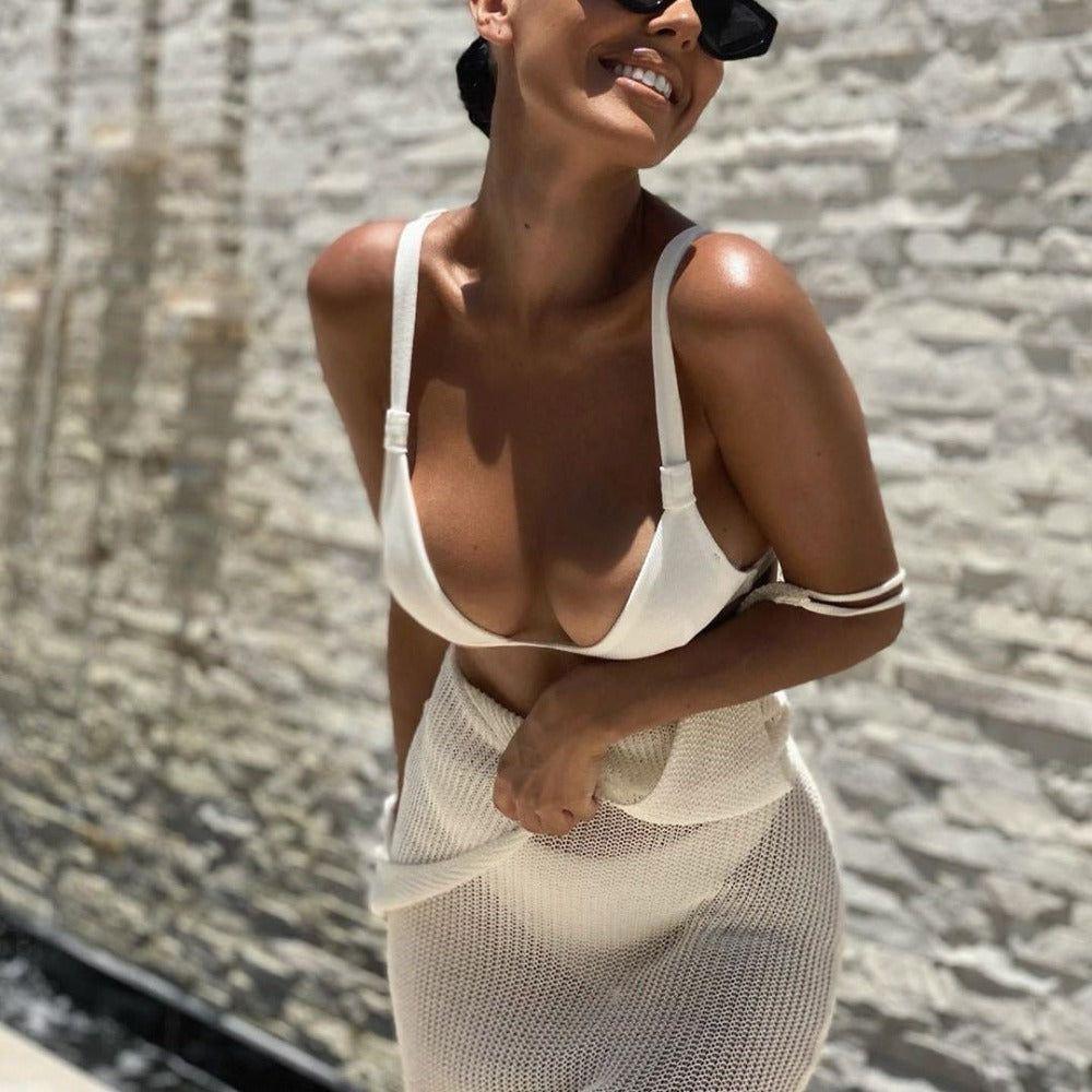 Woman wearing a white bikini and mesh cover-up against a stone wall.