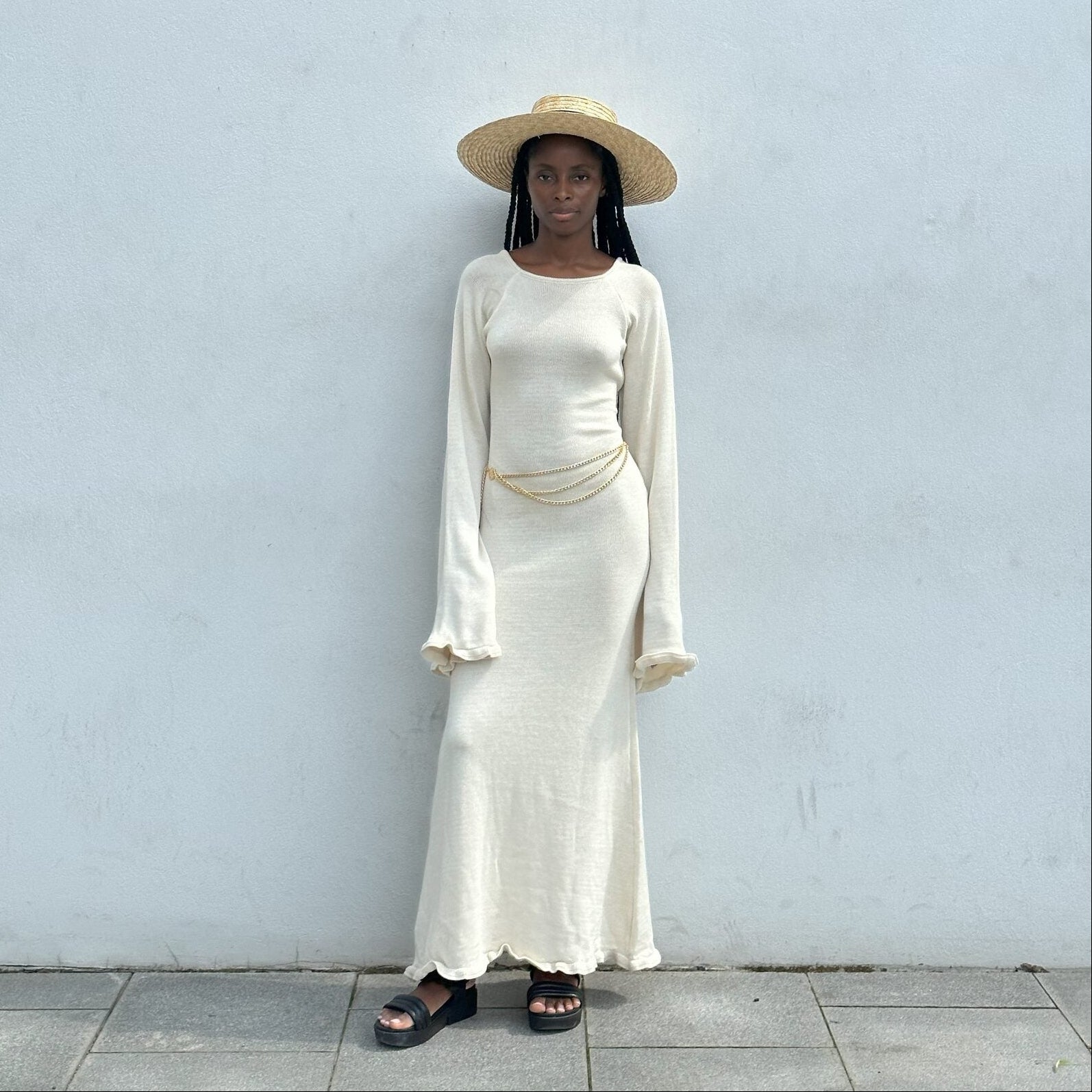 Person wearing a long cream dress and straw hat against a light gray wall.