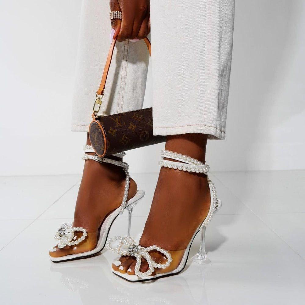 Brown high-heeled shoes with pearl embellishments worn by a person holding a Louis Vuitton handbag.