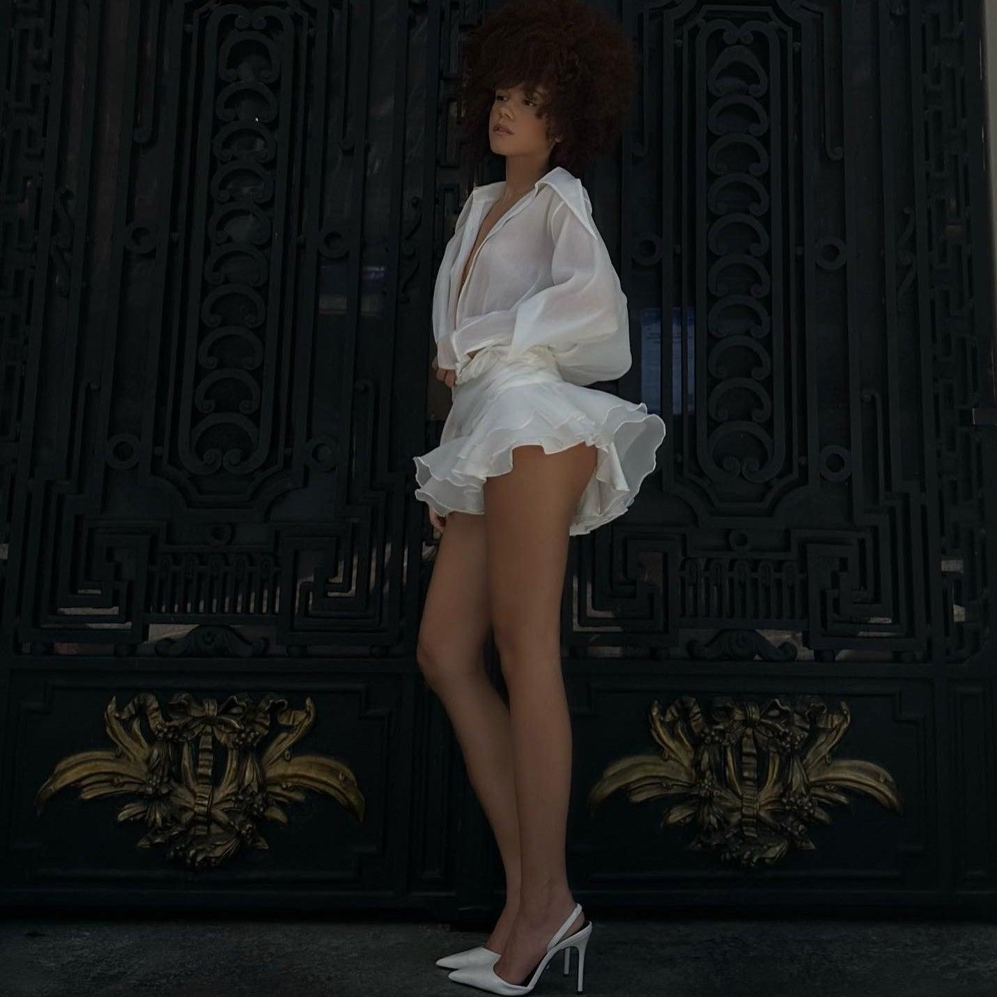 Sheer white blouse with deep neckline and ruffled mini skirt, paired with pointed-toe heels,