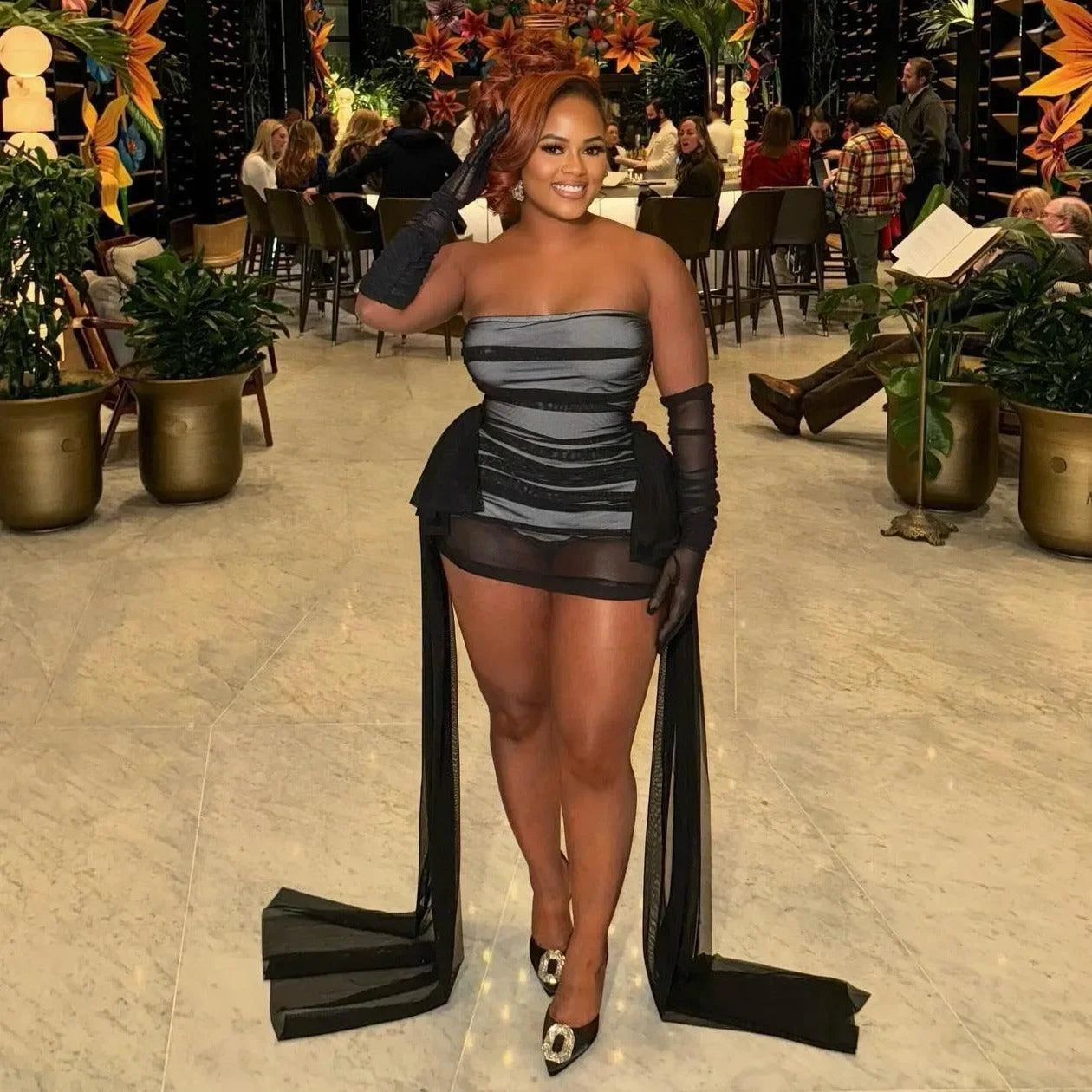 Black mini dress featuring ruched details, strapless design, and dramatic sheer panel accents, styled with gloves for a...