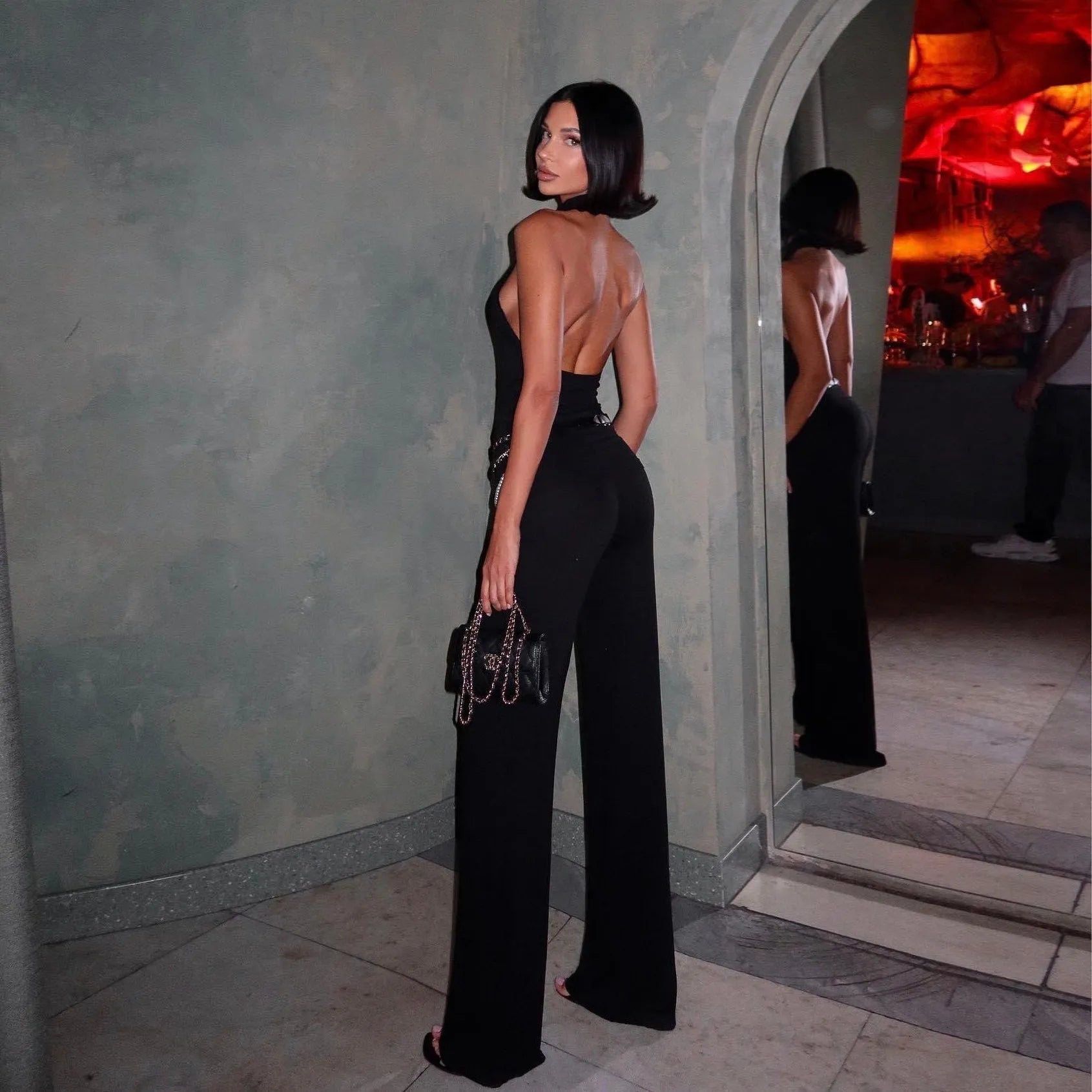Open-back black jumpsuit with wide legs, paired with a chain-strap bag, stylish evening look.