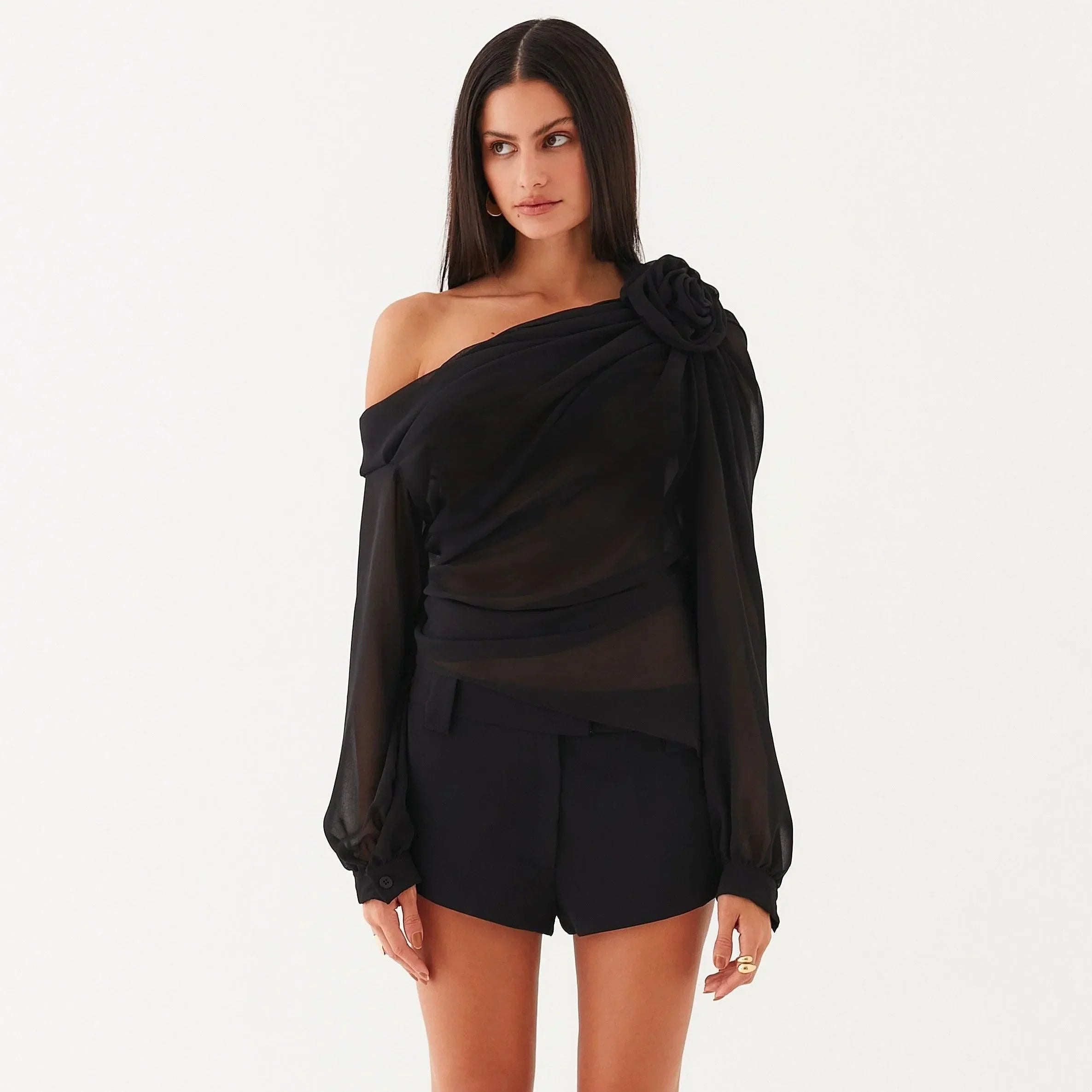 Black asymmetrical draped blouse with floral detail, styled off one shoulder, paired with tailored shorts for a chic look.
