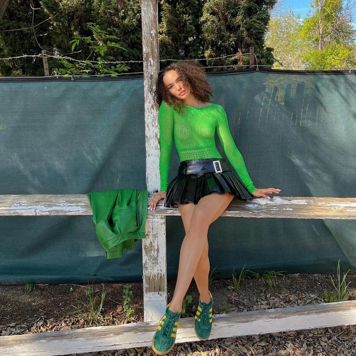 Green mesh backless bodysuit with diamond details, styled with a black pleated skirt and green sneakers for a chic look.