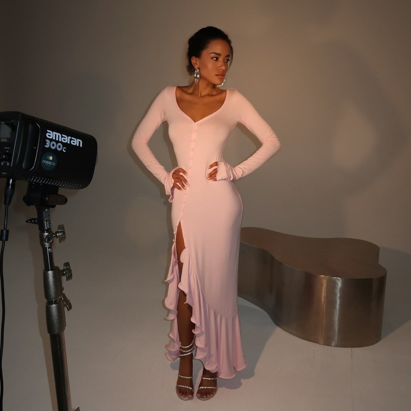 Pink ruffled midi dress with long sleeves, V-neck, front slit, styled with strappy heels for a chic look.