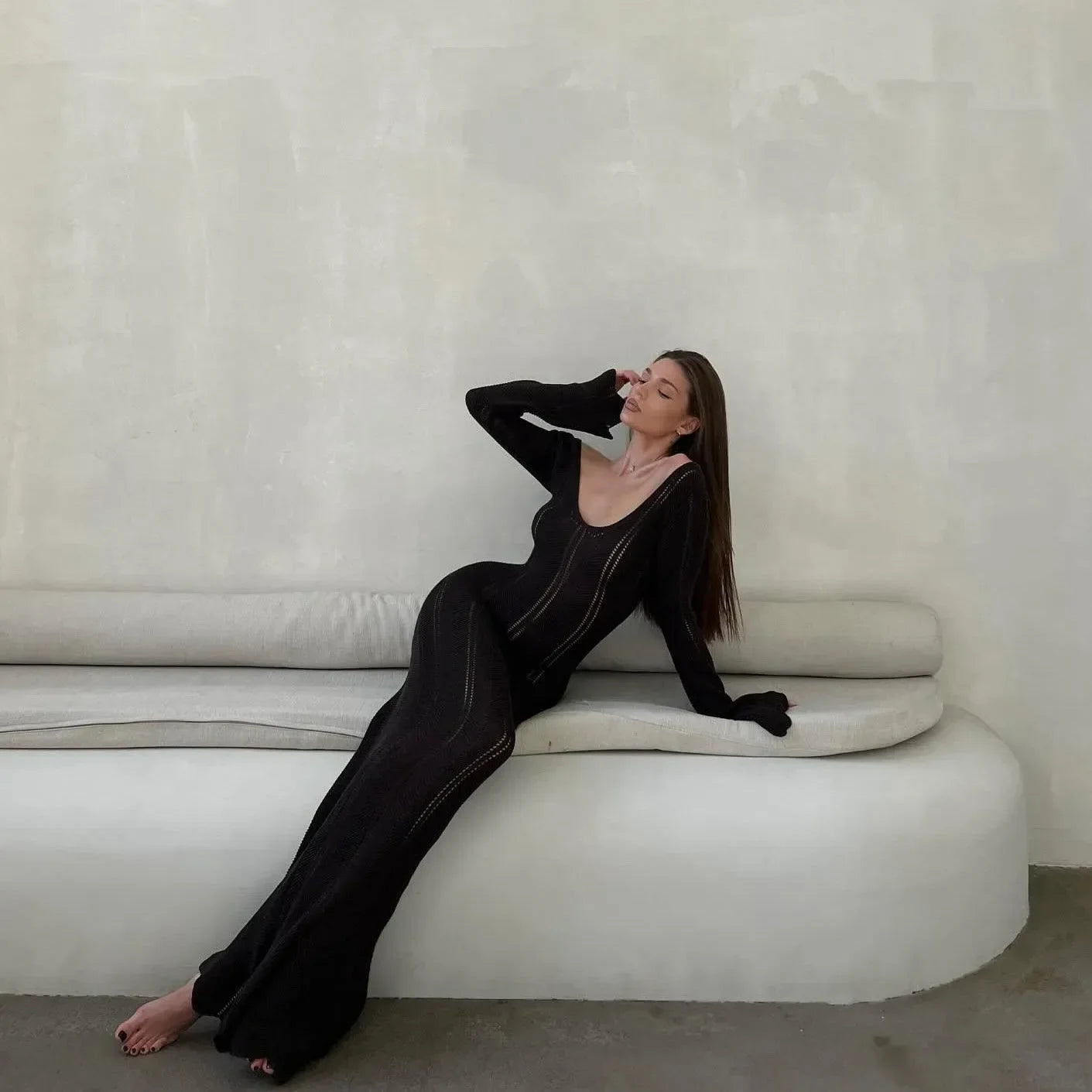 Black long-sleeved maxi dress with a deep V-neck and fitted silhouette, elegantly worn with a relaxed pose on a sofa.