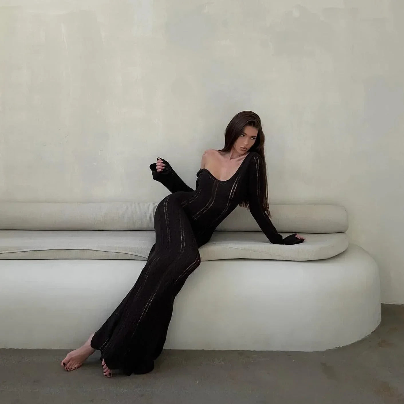 Black knitted long-sleeved maxi dress with a fitted silhouette, styled with one shoulder exposed while lounging.