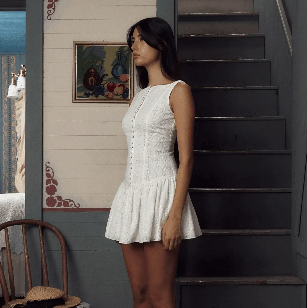 White embroidered mini dress with a fitted bodice, flared skirt, and button detail, styled for a casual summer look.