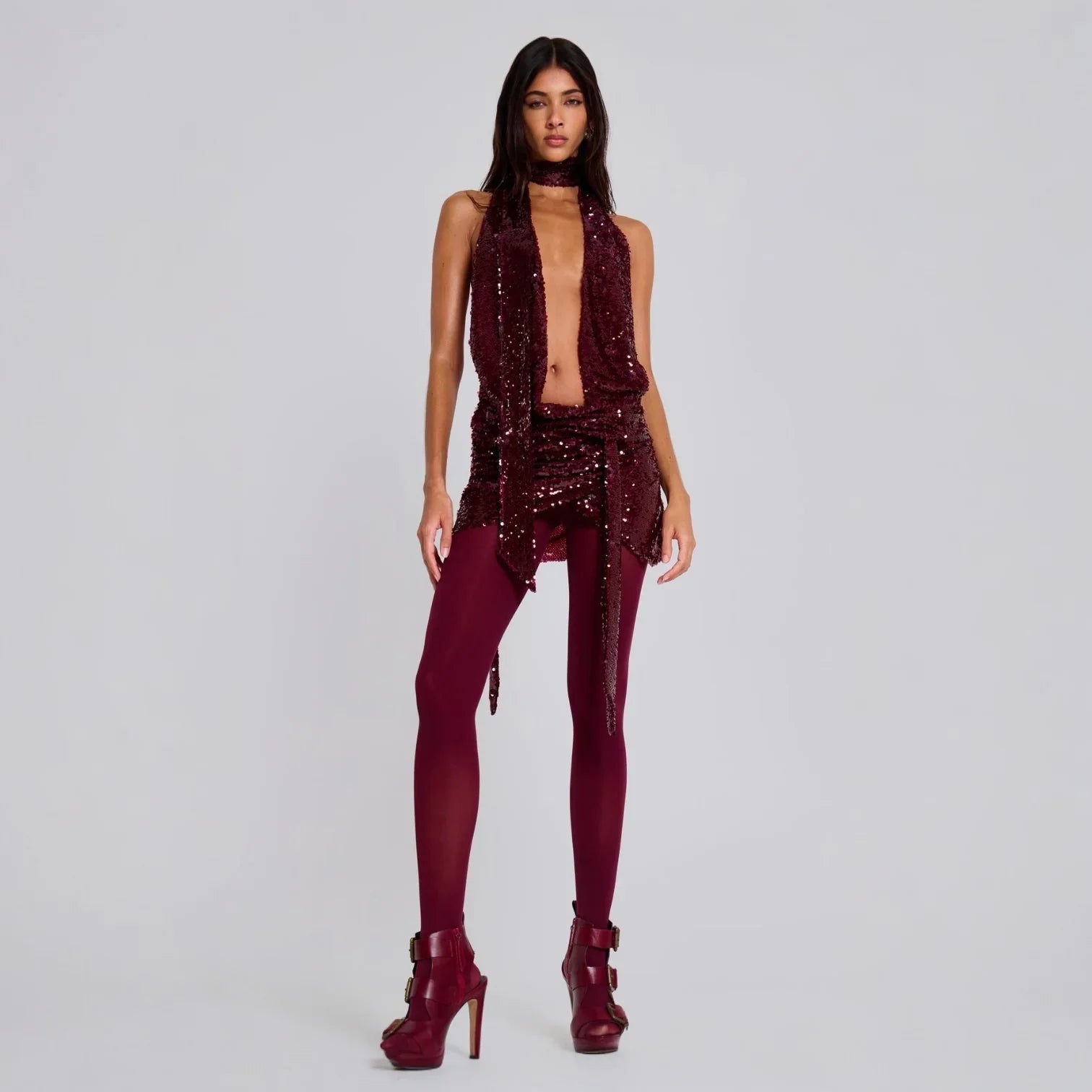 Burgundy backless sequin halterneck mini dress with a tied waist, styled with matching leggings and heels.