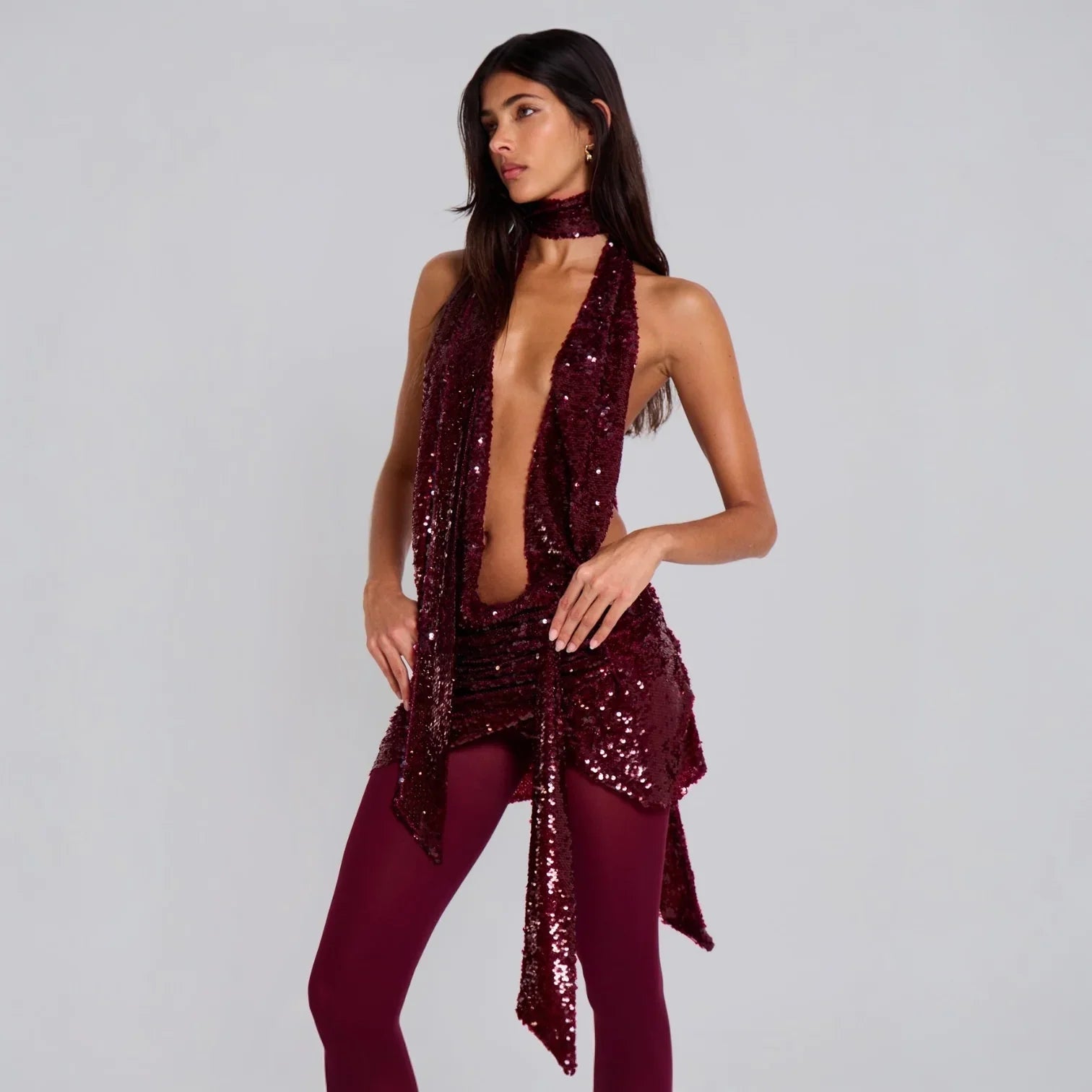Backless burgundy sequin halterneck mini dress with draped front and neck tie, styled with fitted leggings.