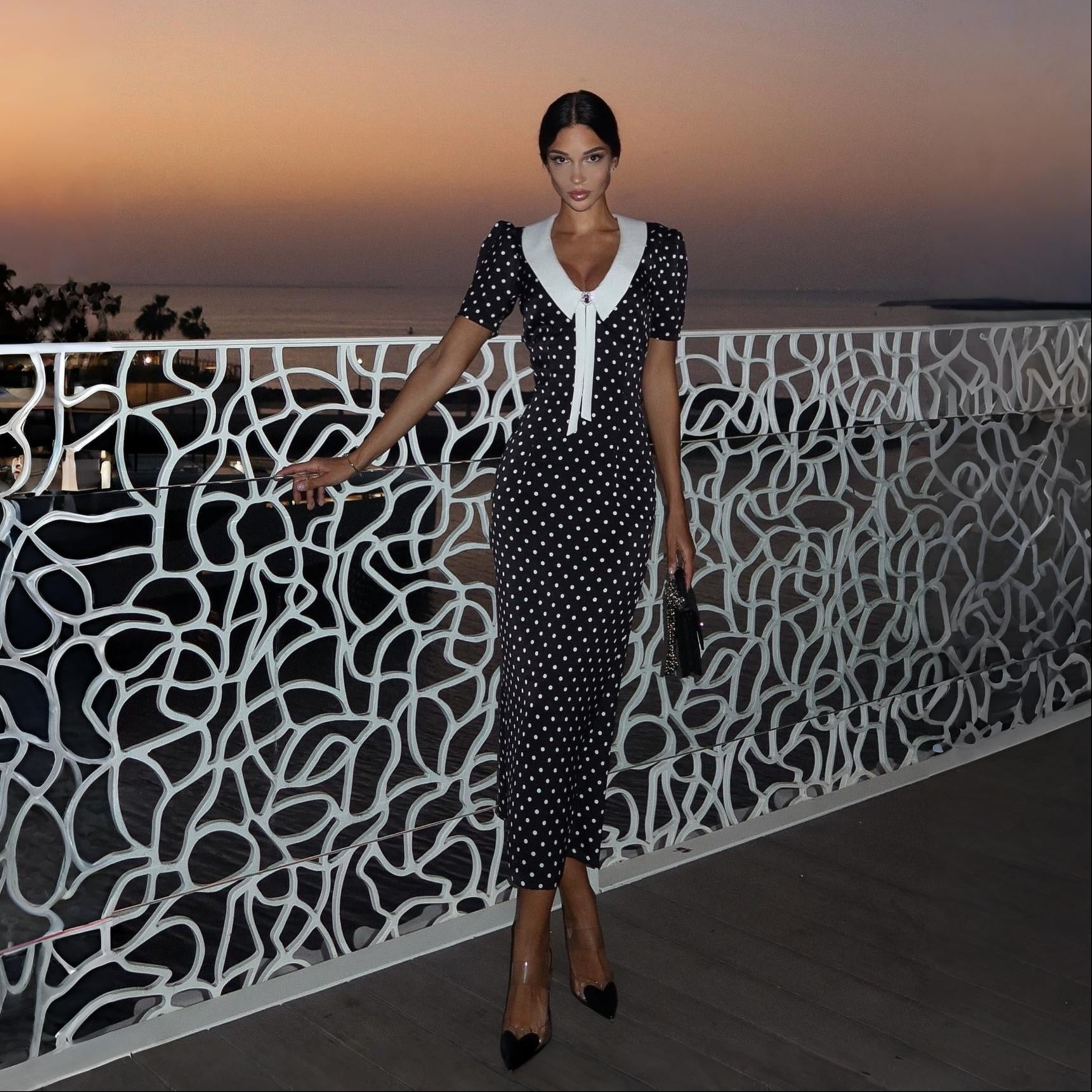 Black polka dot midi dress with a white collar, fitted silhouette, and puff sleeves, styled with heels and a clutch.