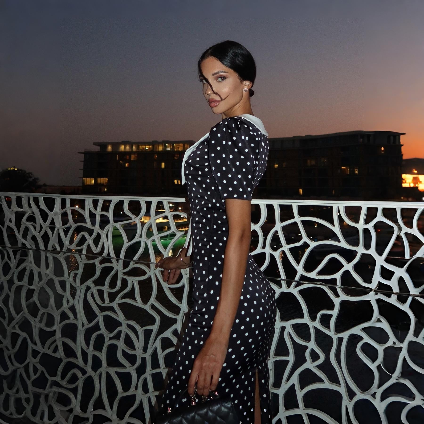 Black polka dot midi dress with a white collar, fitted silhouette, styled for an evening look against a sunset backdrop.