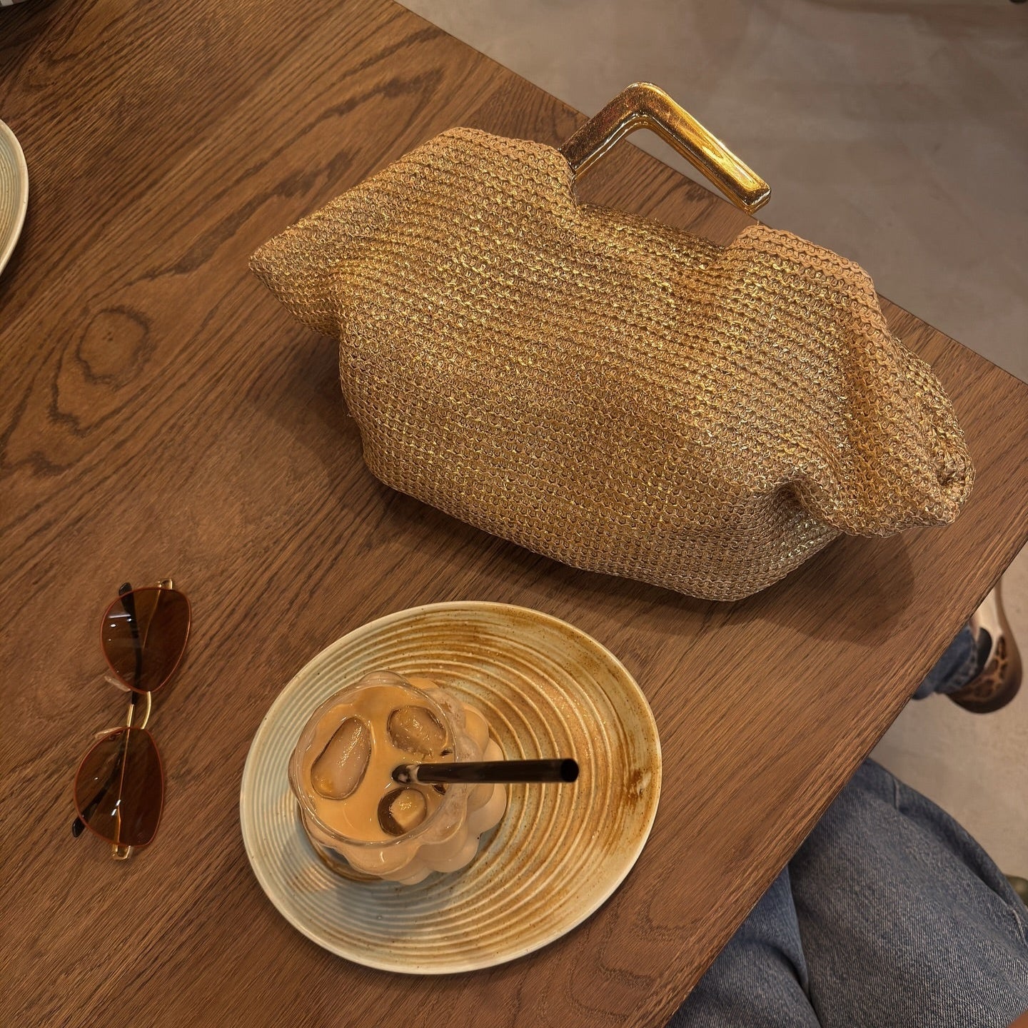 Gold woven handbag with metal handle, styled casually on a wooden table next to sunglasses and an iced drink.