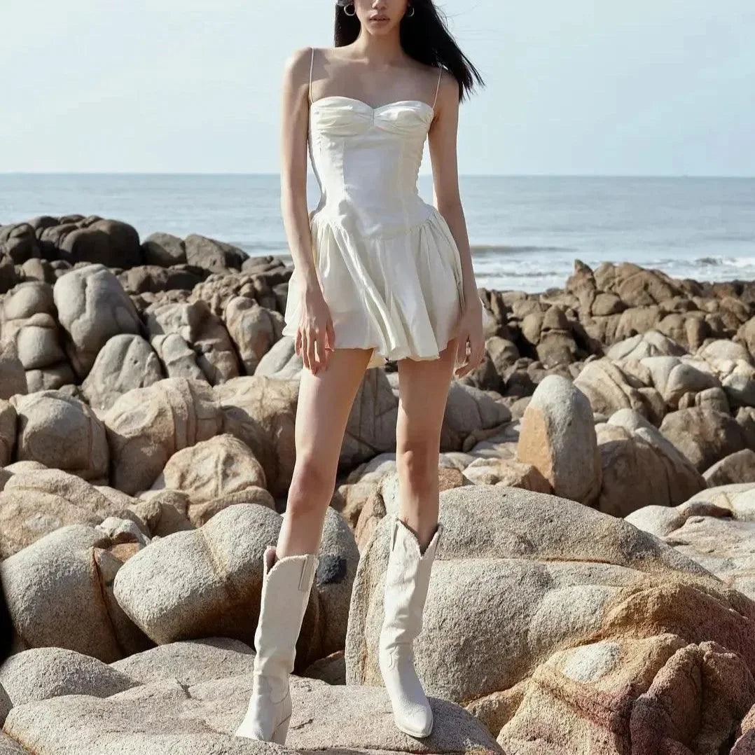 Light beige mini dress with fitted bodice and flared skirt, styled with knee-high white boots on rocky beach.