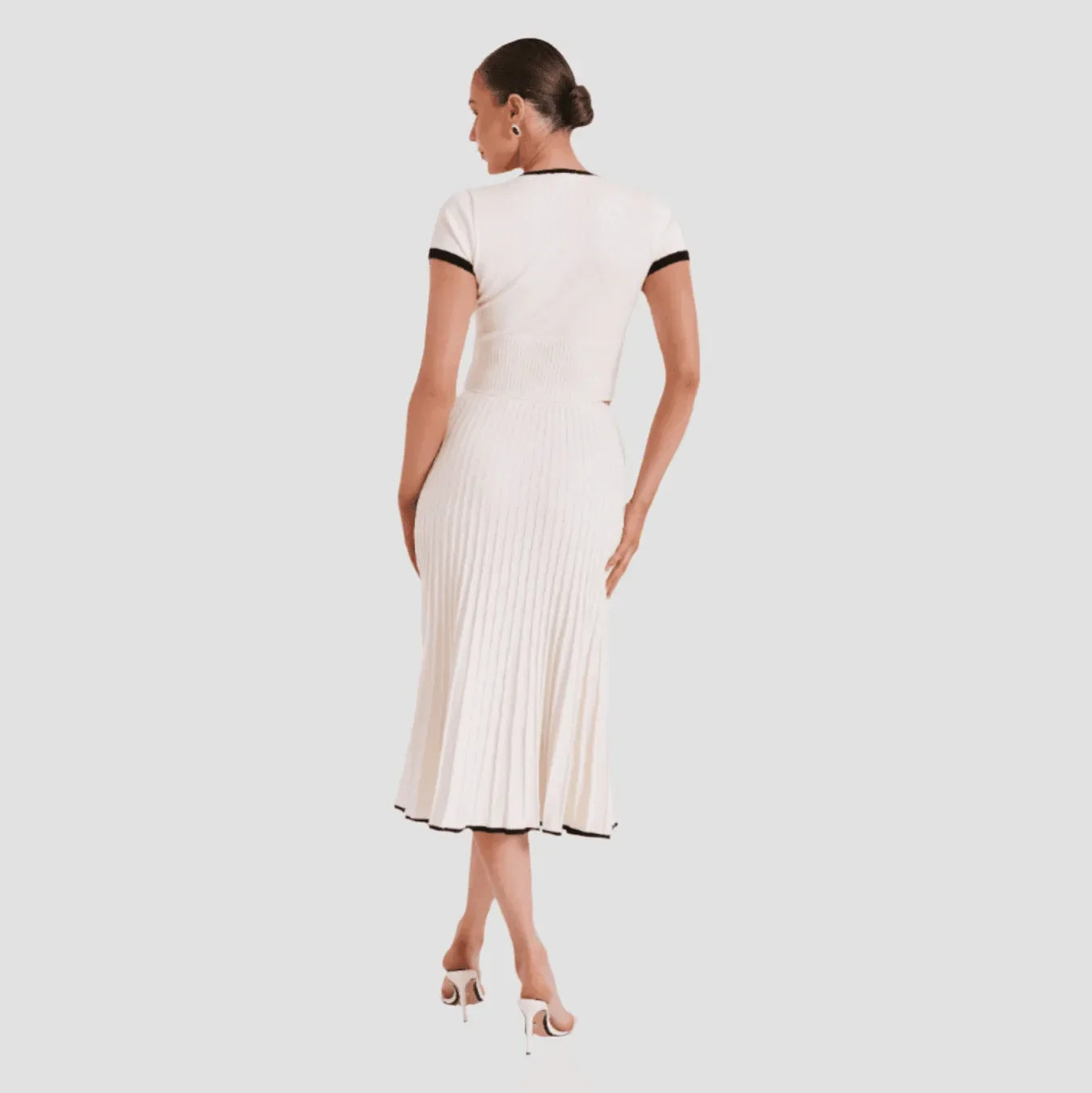 Cream two-piece set featuring a fitted top with black trim and a pleated midi skirt, styled with strappy heels.