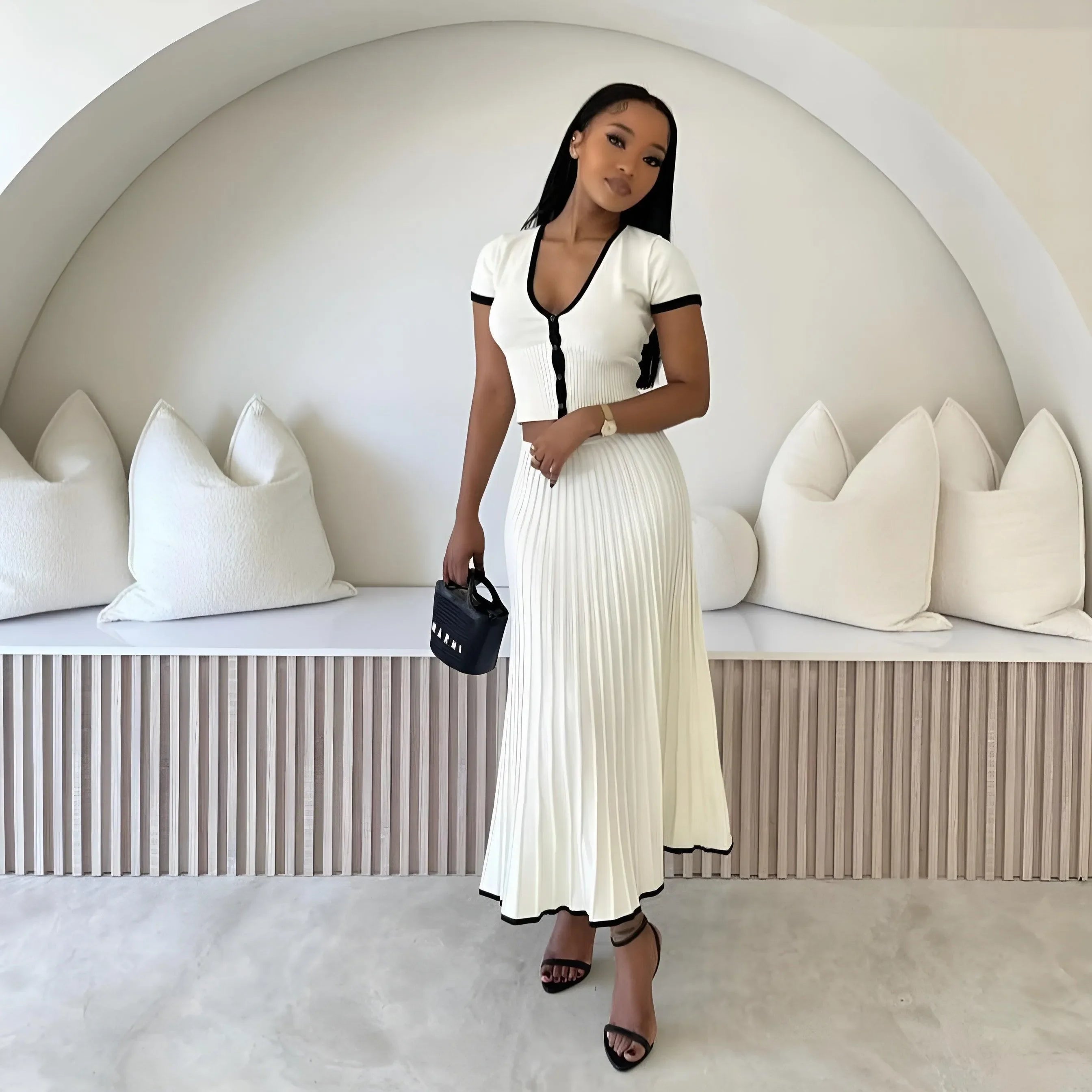 White two-piece set featuring a fitted short-sleeve cardigan with black trim and a pleated midi skirt, styled with...