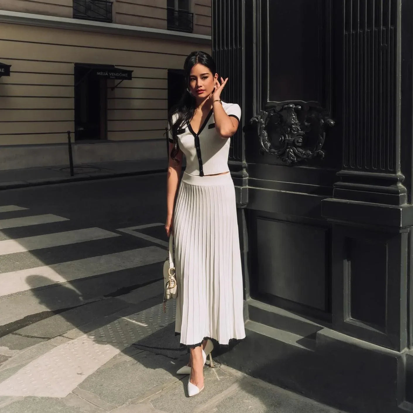 Chic white contrast knit two-piece set with a fitted top and pleated skirt, styled with heeled pumps and a clutch.