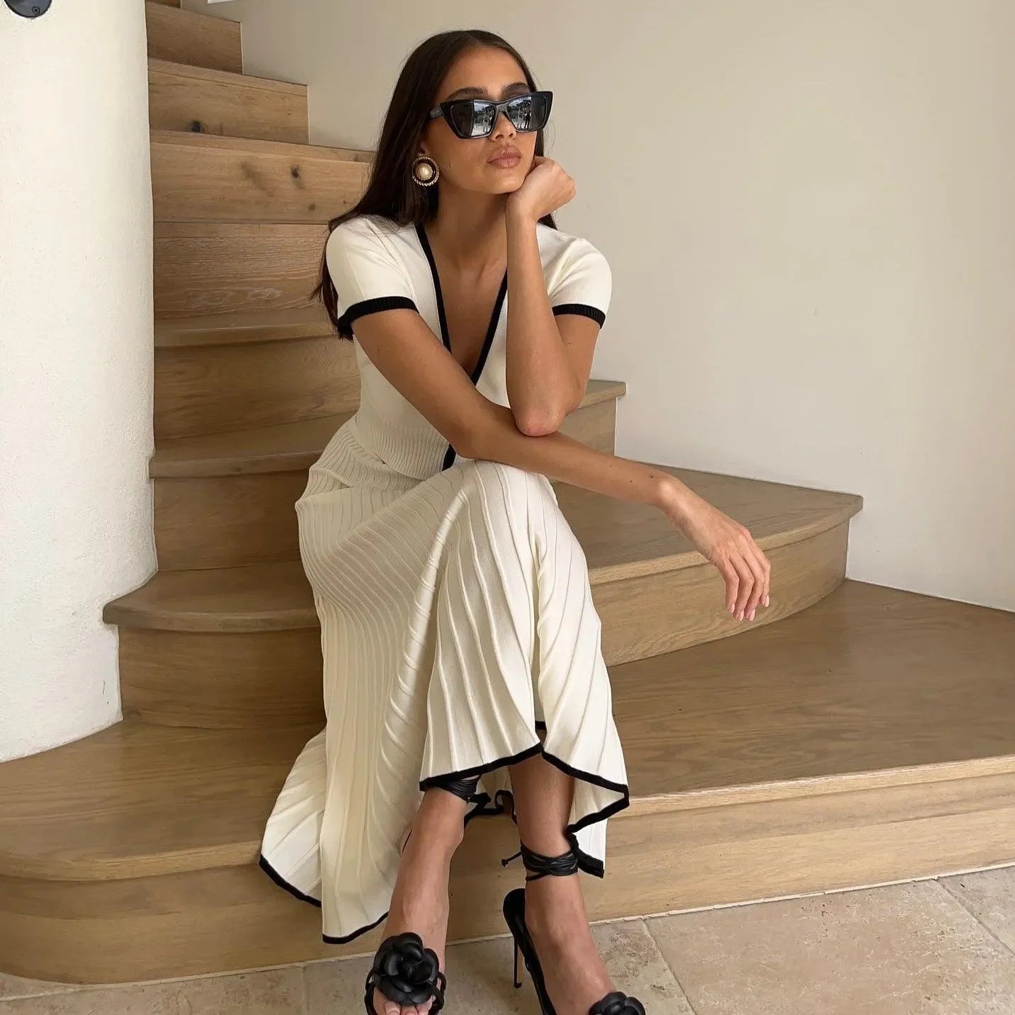 Cream two-piece set with black trim, featuring a fitted top and flowing pleated skirt, styled with sunglasses and heels.