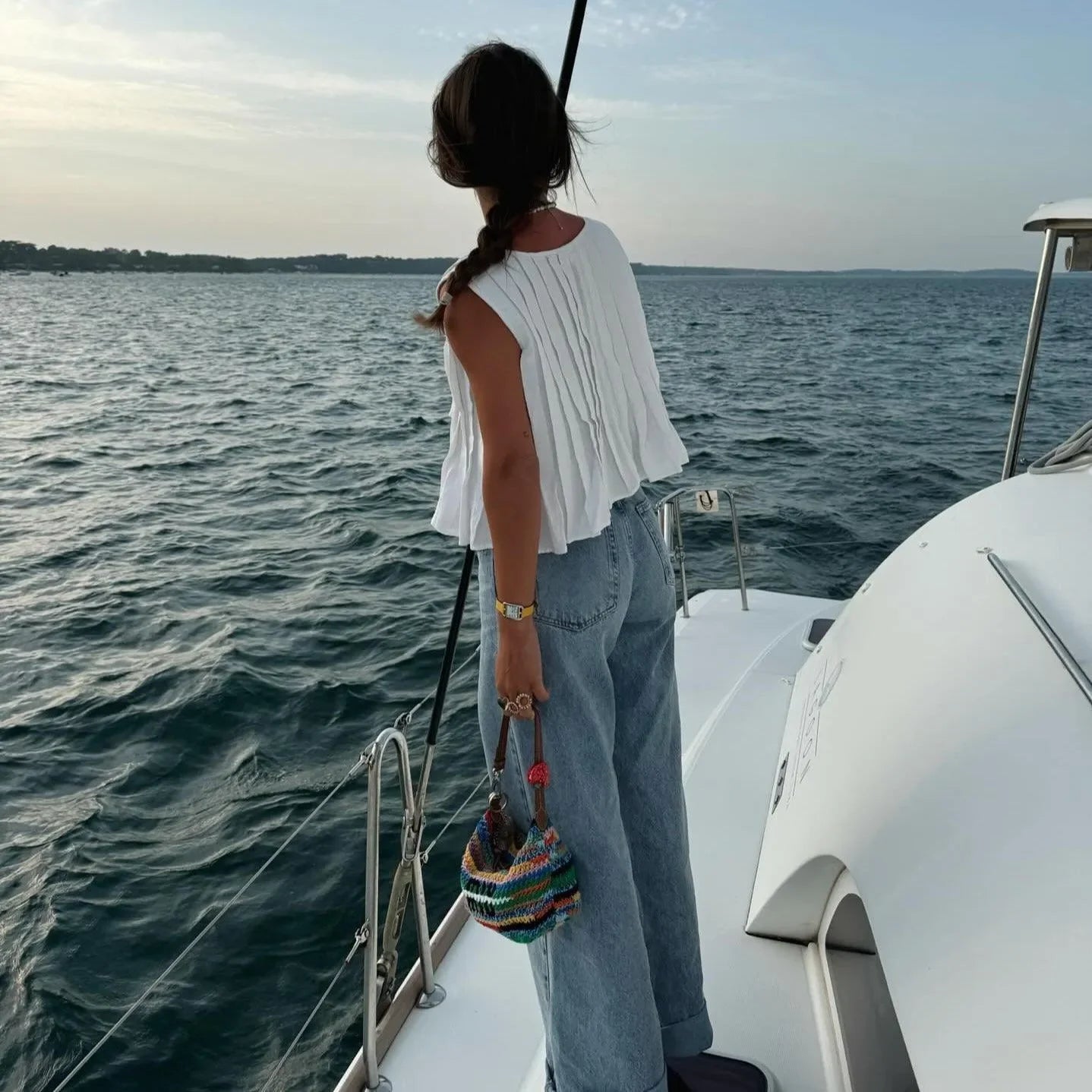 White pleated cotton top paired with light blue high-waisted jeans, styled with a colorful handbag on a boat.