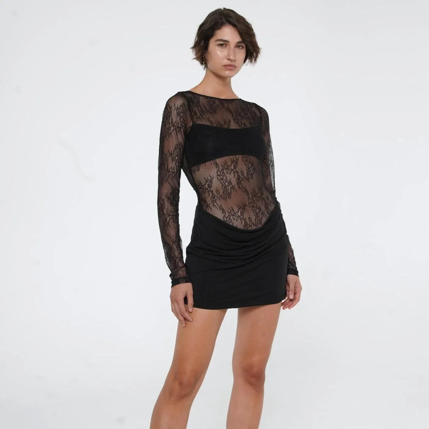Sheer black lace mini dress with long sleeves and ruched skirt, styled for a chic, playful evening look.