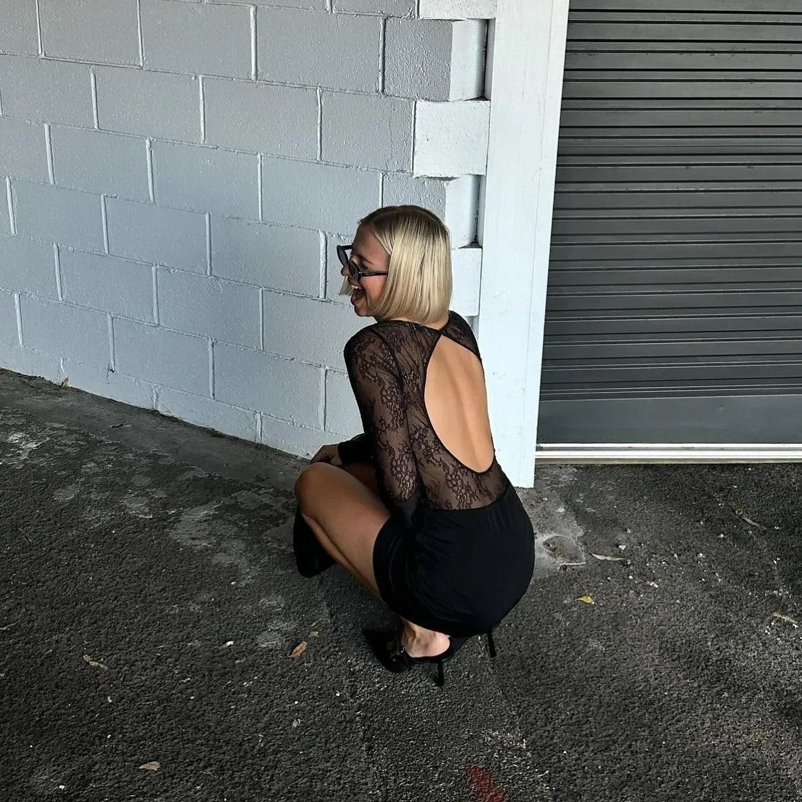 Black lace mini dress with a backless design, styled casually with sunglasses and heels for a chic look.