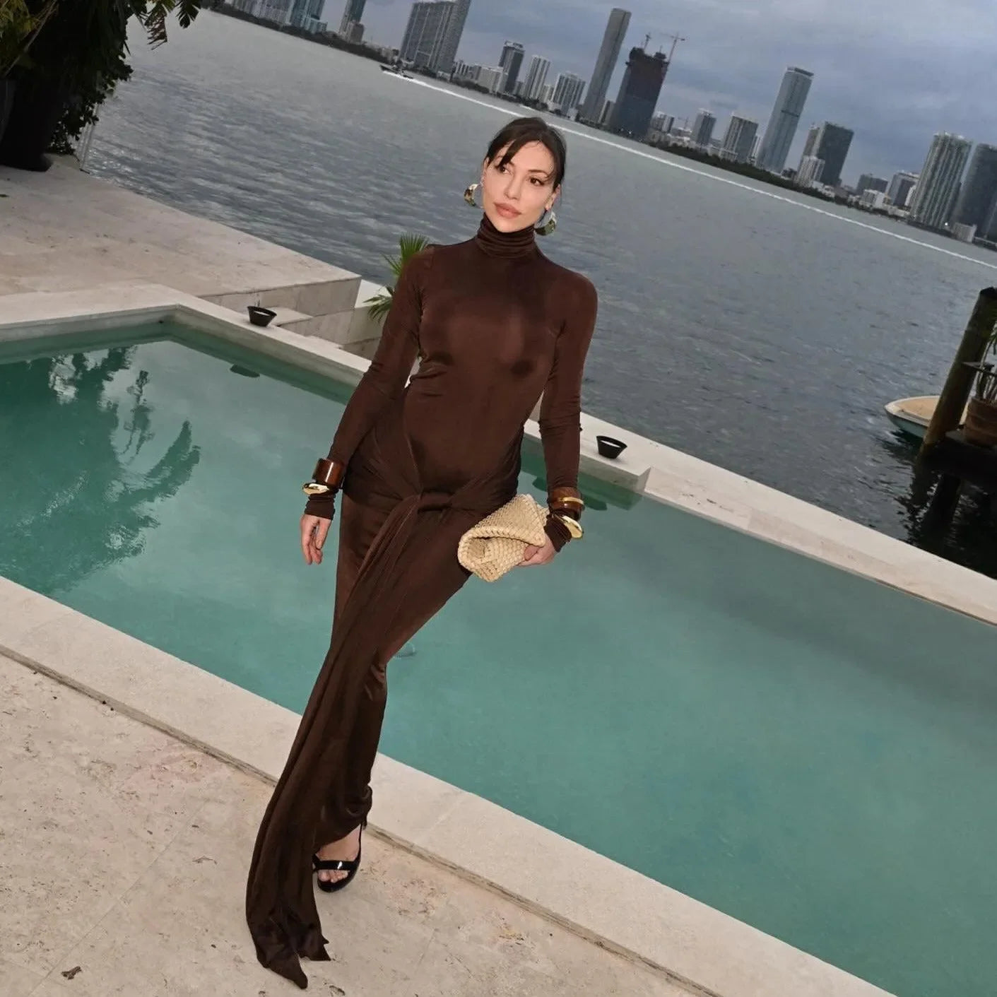 Brown long sleeve maxi dress with a high neckline and side slit, styled with elegant accessories and heels.