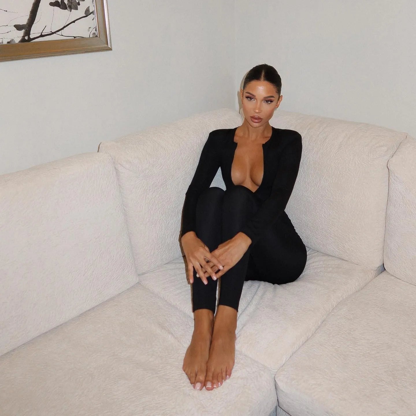 Black long sleeve jumpsuit featuring a deep V-neck, styled casually with legs crossed on a sofa. Perfect for chic comfort.