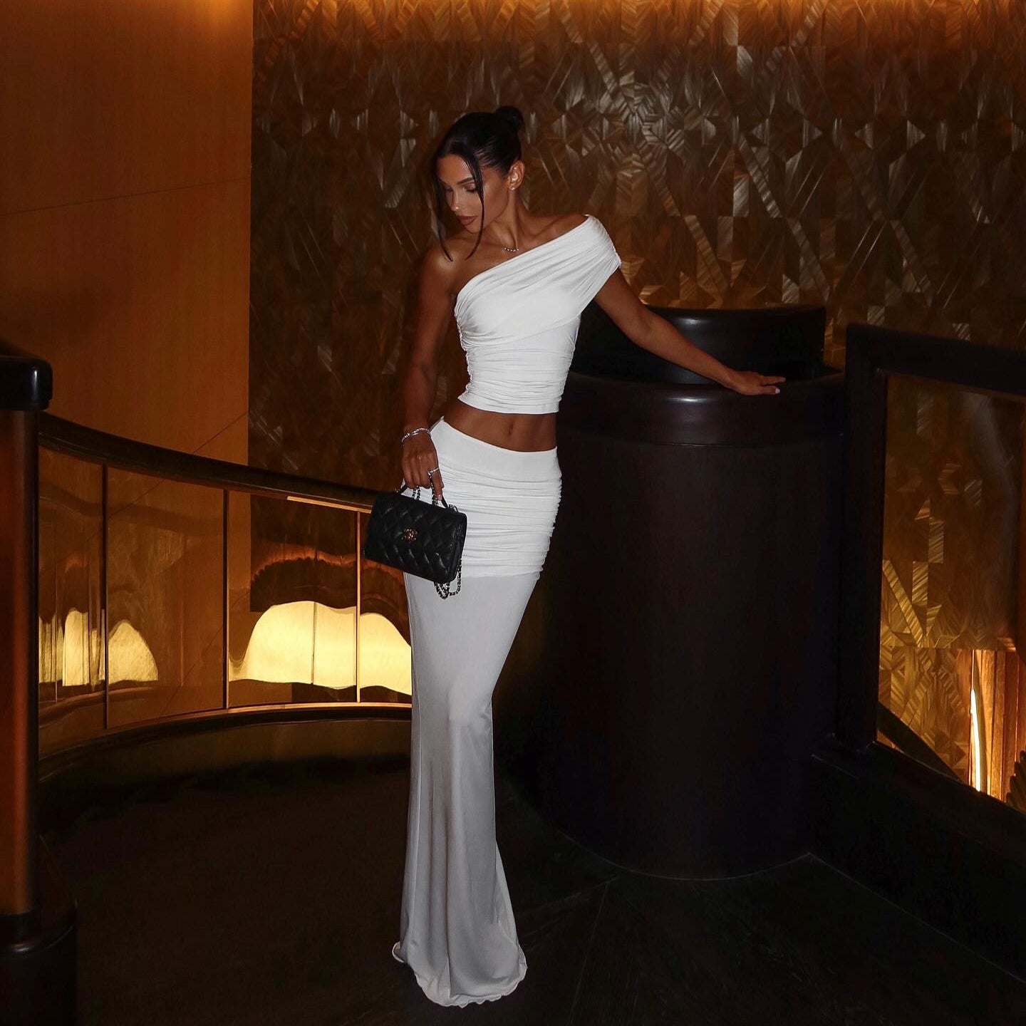White one-shoulder two-piece set with ruched detailing, styled with a chic black purse and sleek hair for an elegant look.