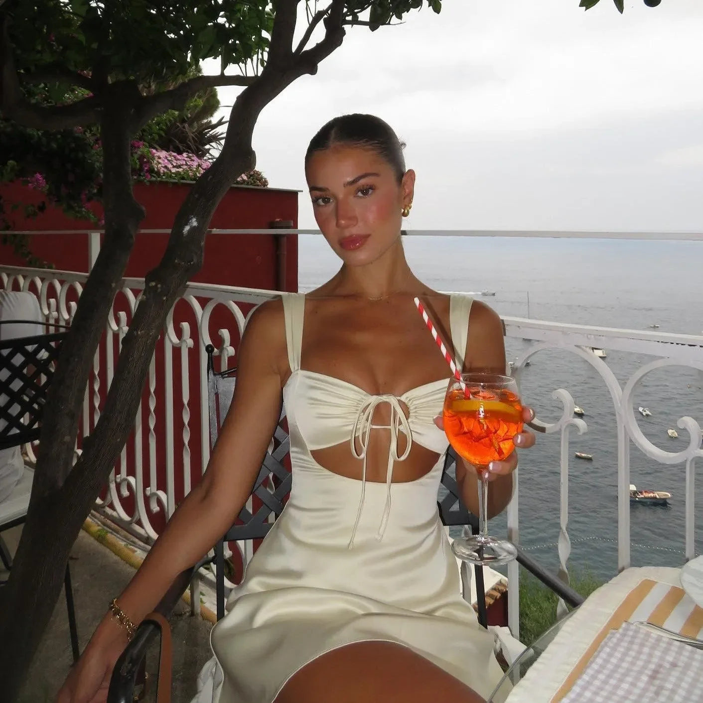 Ivory mini dress in satin with tie-front and cut-out details, styled with a drink for a chic summer look.